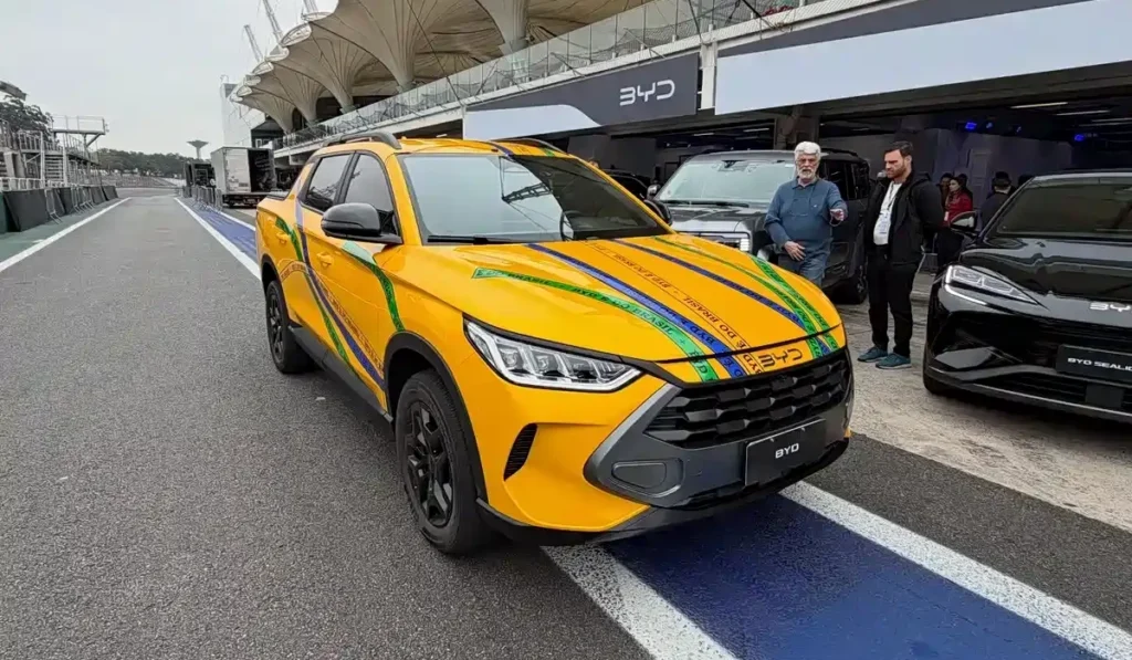 Yellow BYD pickup/SUV with blue and green racing stripes parked on a race track pit lane, front view with a black grille and 'BYD' badge visible.