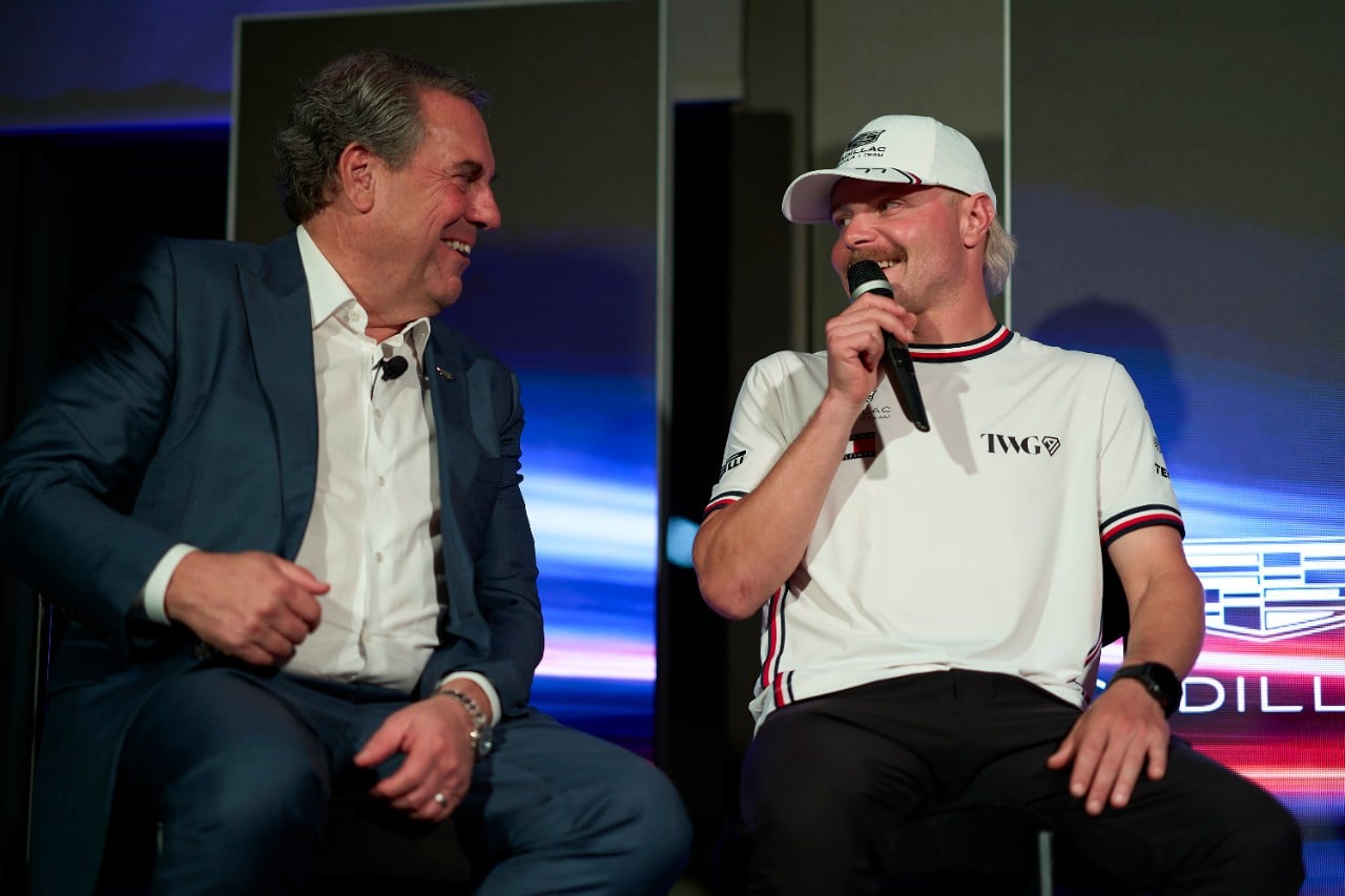 Two men sit on a stage, one in a blue suit smiling at the other who wears a white cap and shirt, holding a microphone.