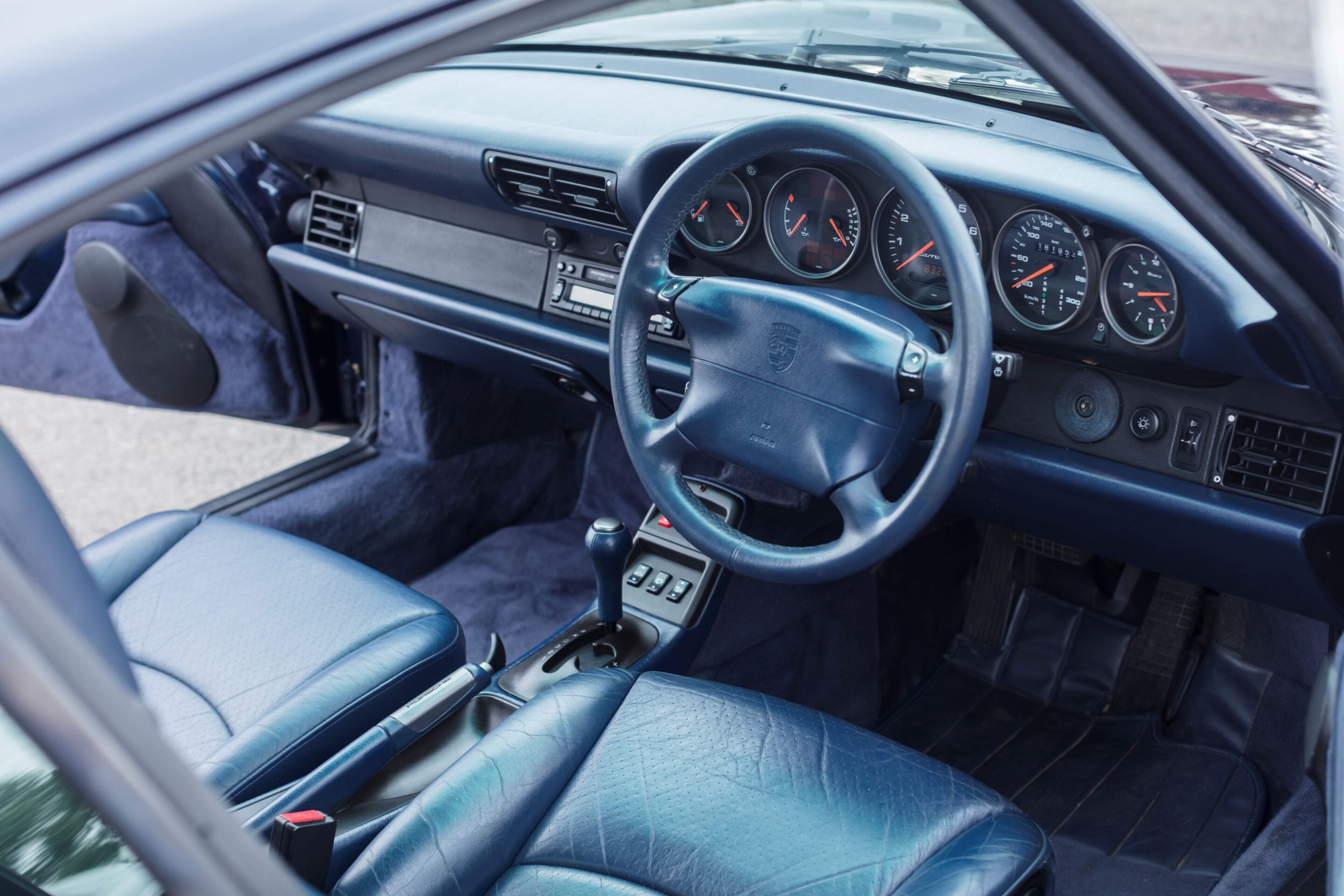 Blue leather car interior with a multi-gauge dashboard and steering wheel seen from the passenger side edge; close-up of driver area including gear shifter and center console.