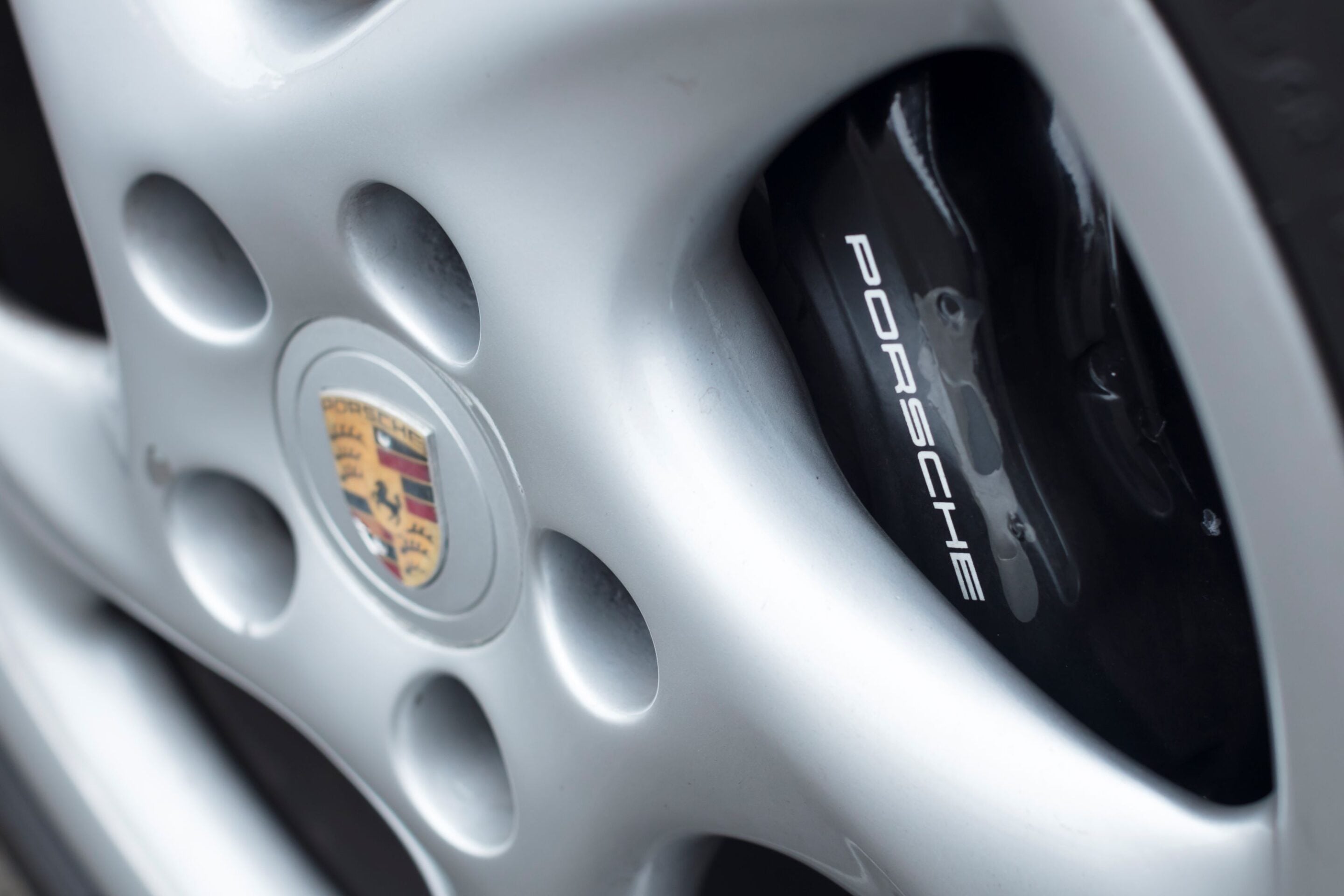 Close-up of a silver Porsche wheel, showing the black brake caliper with 'PORSCHE' branding and the Porsche crest on the hub.