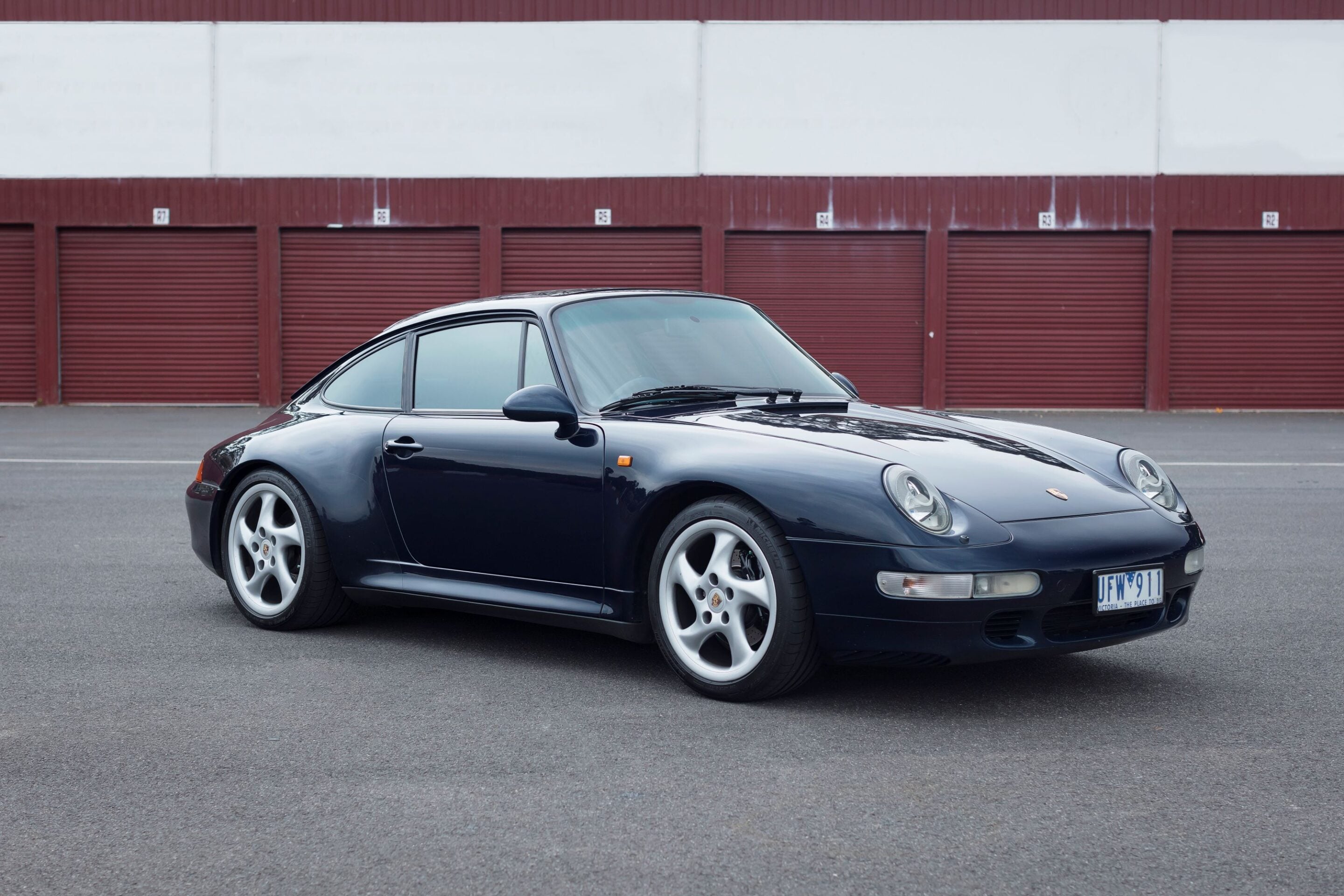 Dark blue Porsche 911 parked on a paved lot in front of red storage unit doors, visible license plate.