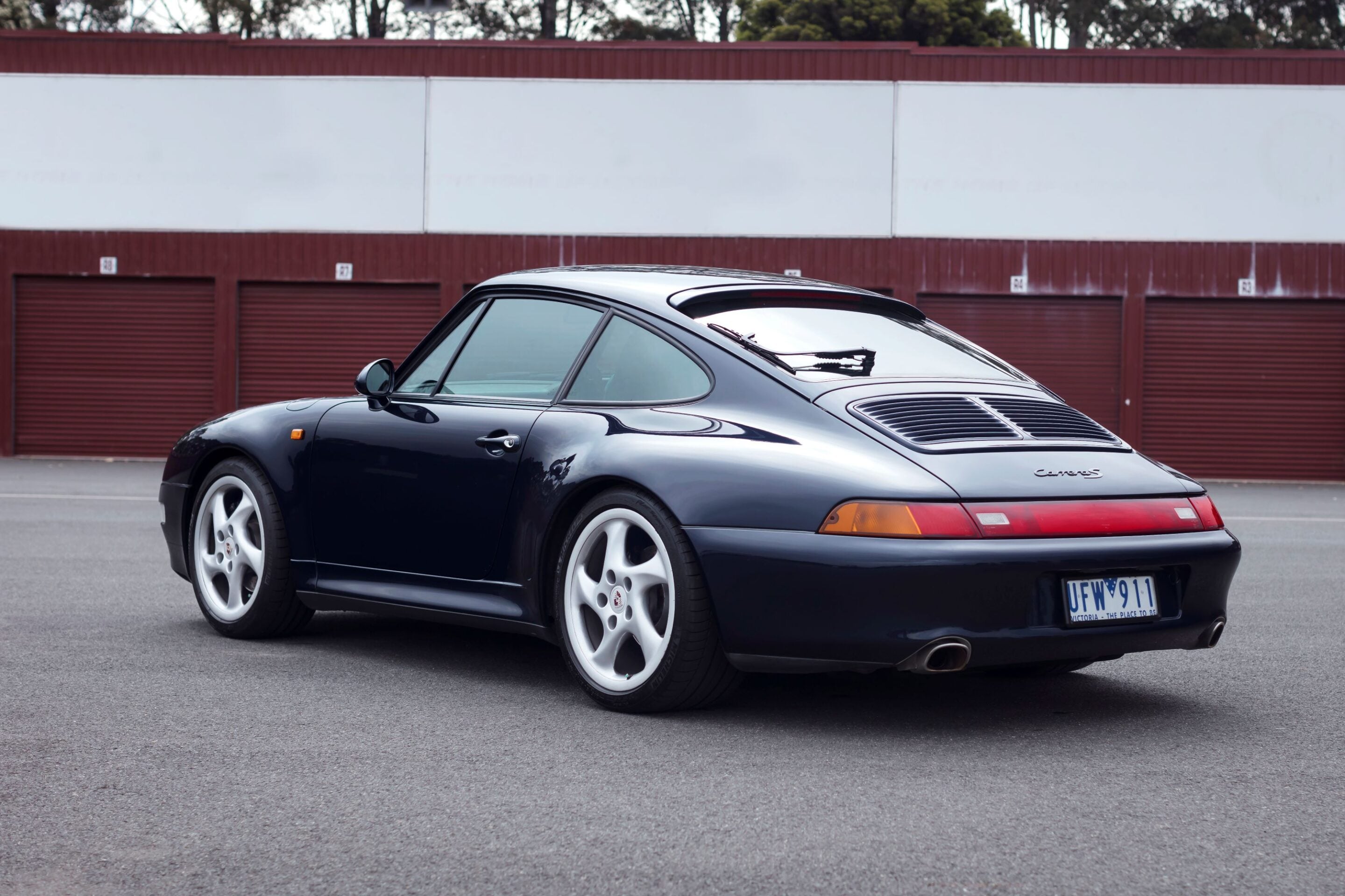 Rear-side view of a dark blue Porsche 911 Carrera S parked in an empty lot with brown garage doors in the background