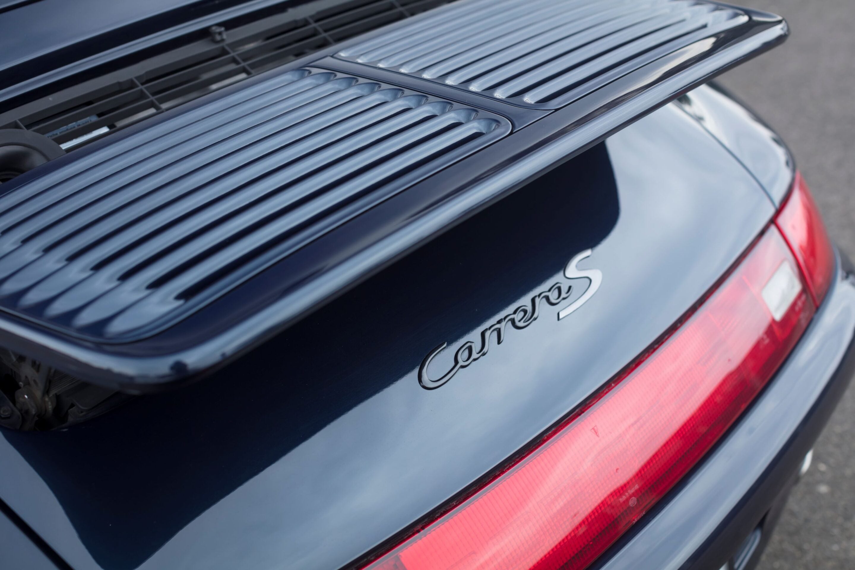 Rear of a dark blue Porsche Carrera S, showcasing the slatted rear spoiler and red taillight section