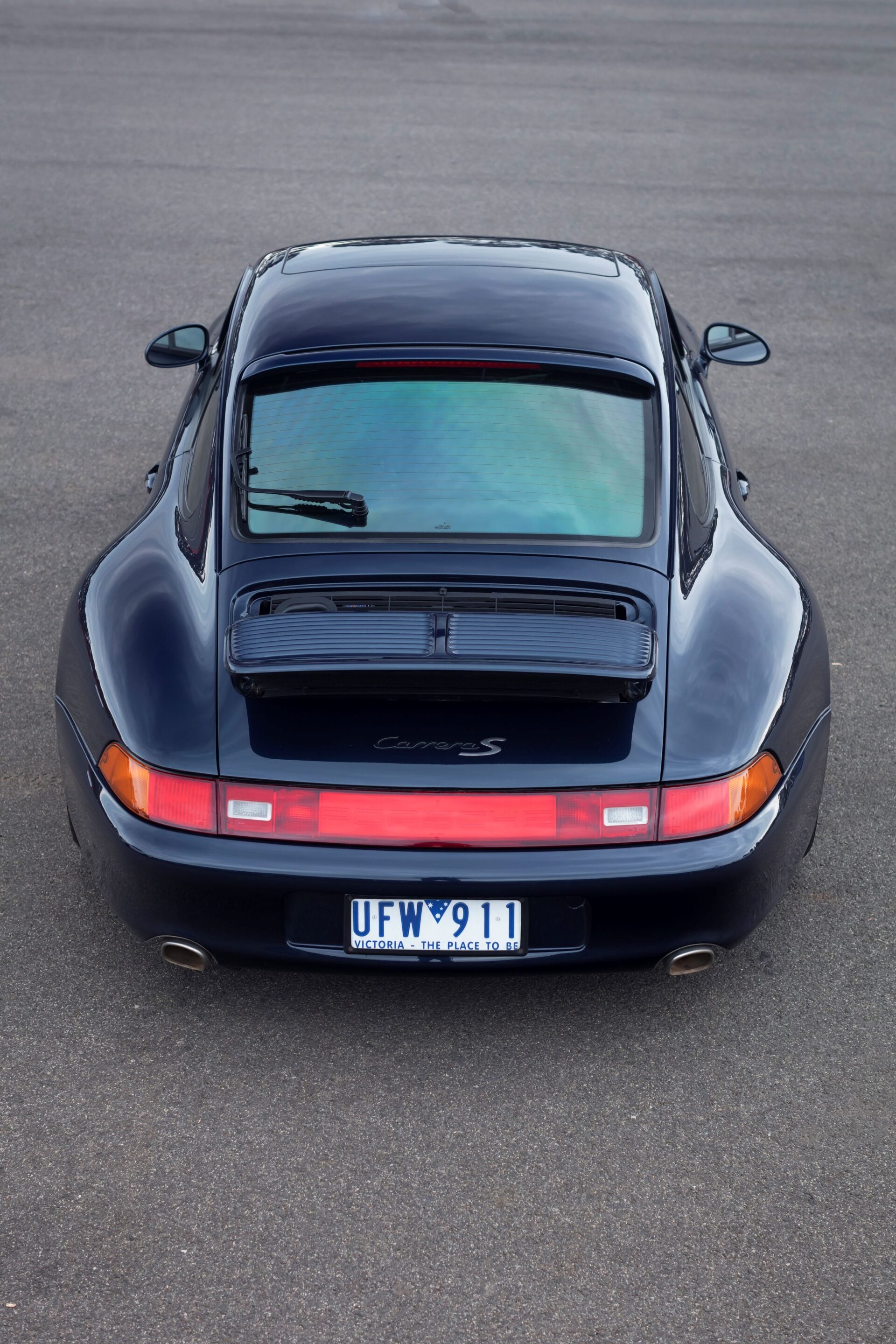 Top-down view of a dark blue Porsche 911 Carrera S parked on asphalt, showing the rear engine cover, rear window, and red taillight strip on either side of the bumper.