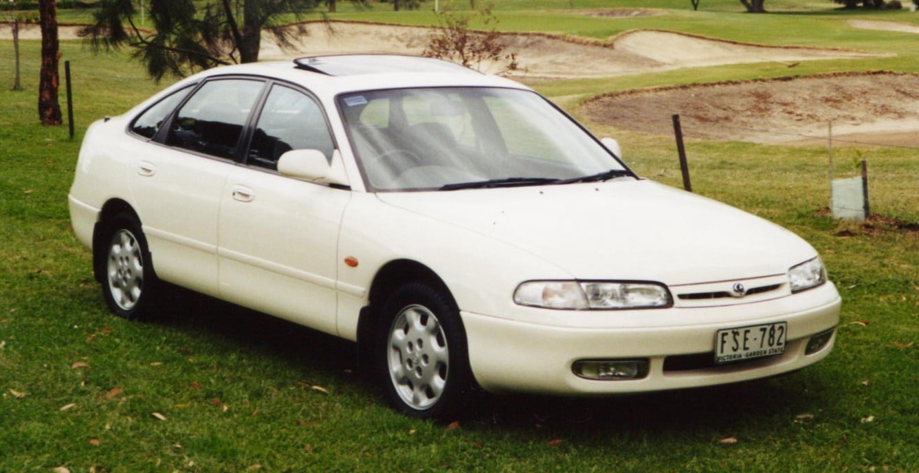 White four-door sedan parked on a grassy field with a golf course in the background, front angled toward the camera, license plate visible but not legible clearly on the bumper.