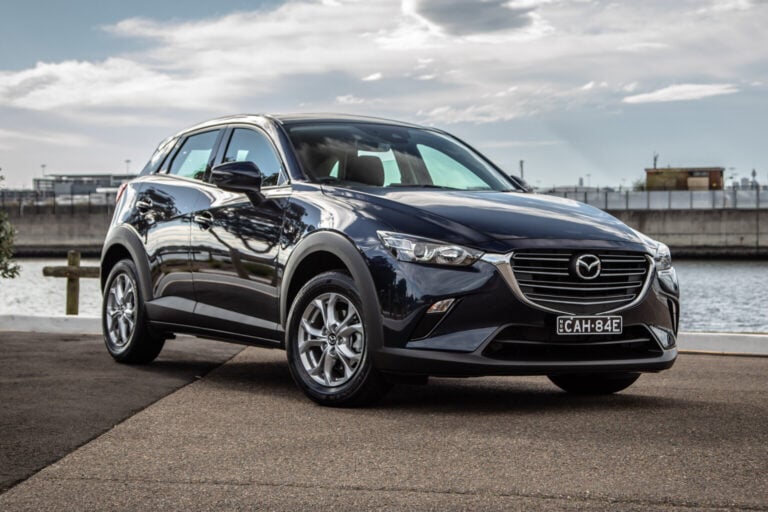 Front three-quarter view of a dark-blue Mazda SUV parked on a waterfront promenade with water and railings in the background