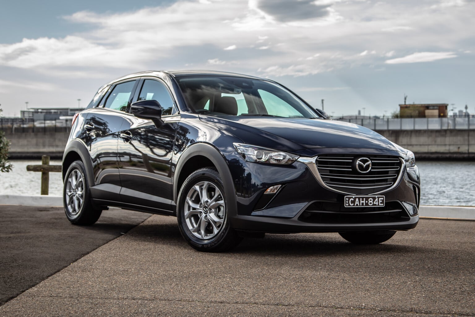 Front three-quarter view of a dark-blue Mazda SUV parked on a waterfront promenade with water and railings in the background