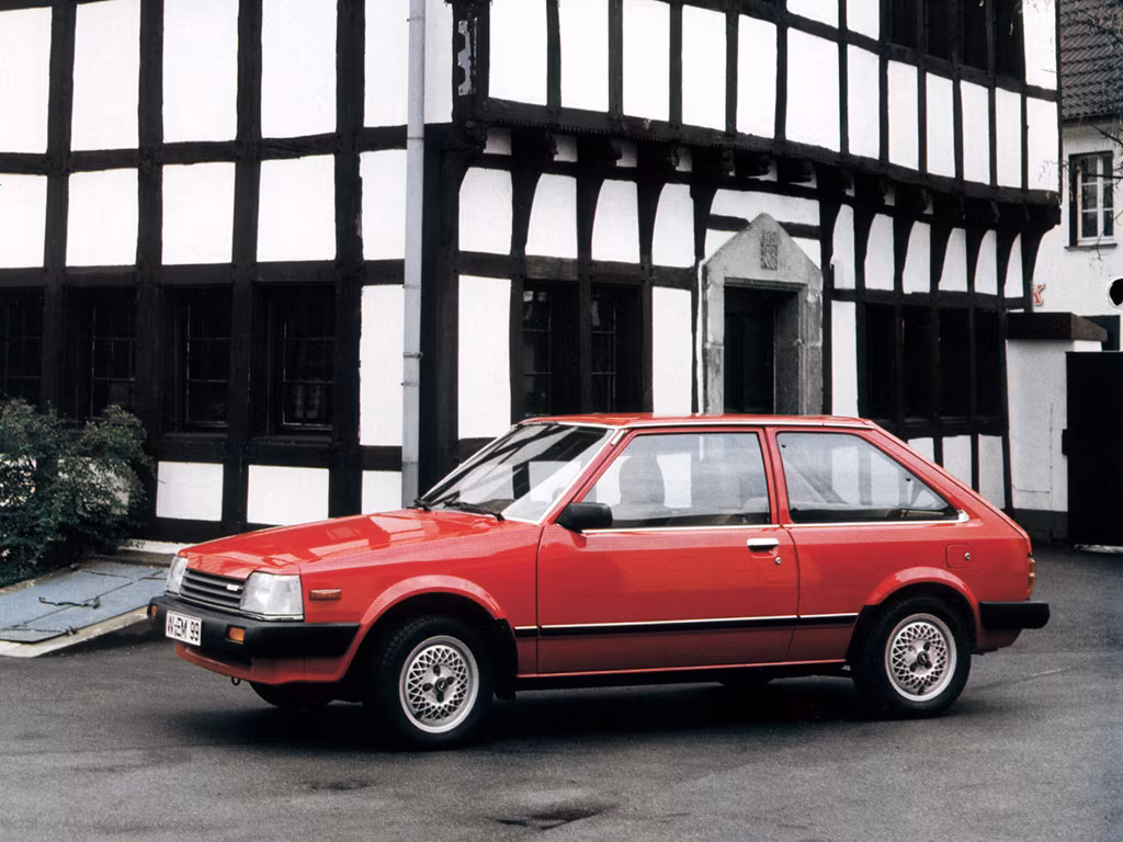 Red vintage hatchback car parked on a street in front of a black-and-white timber-framed building.