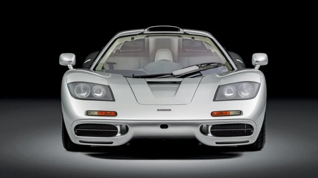 Front view of a silver McLaren sports car against a black backdrop, low, aerodynamic hood visible.