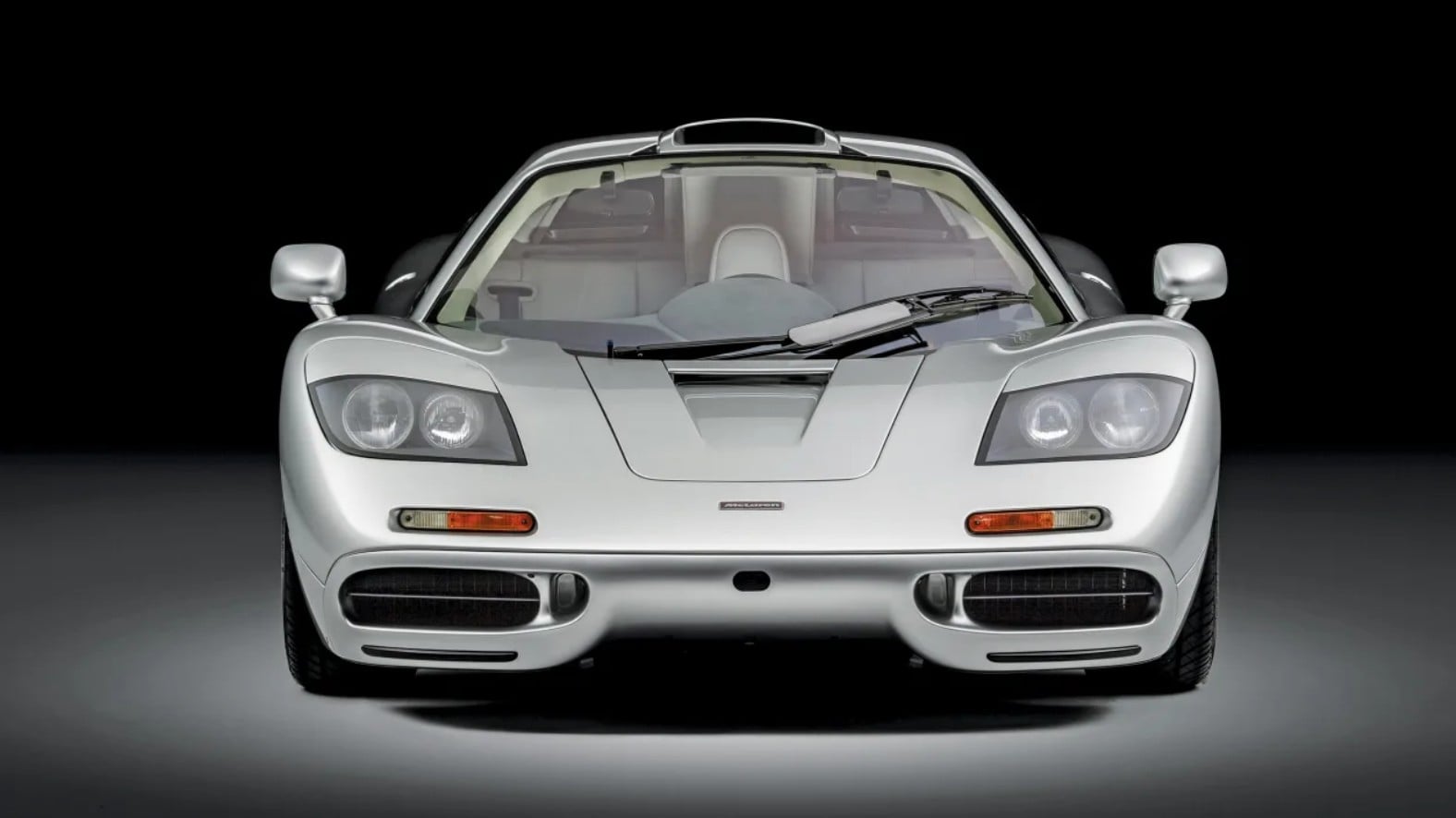 Front view of a silver McLaren sports car against a black backdrop, low, aerodynamic hood visible.