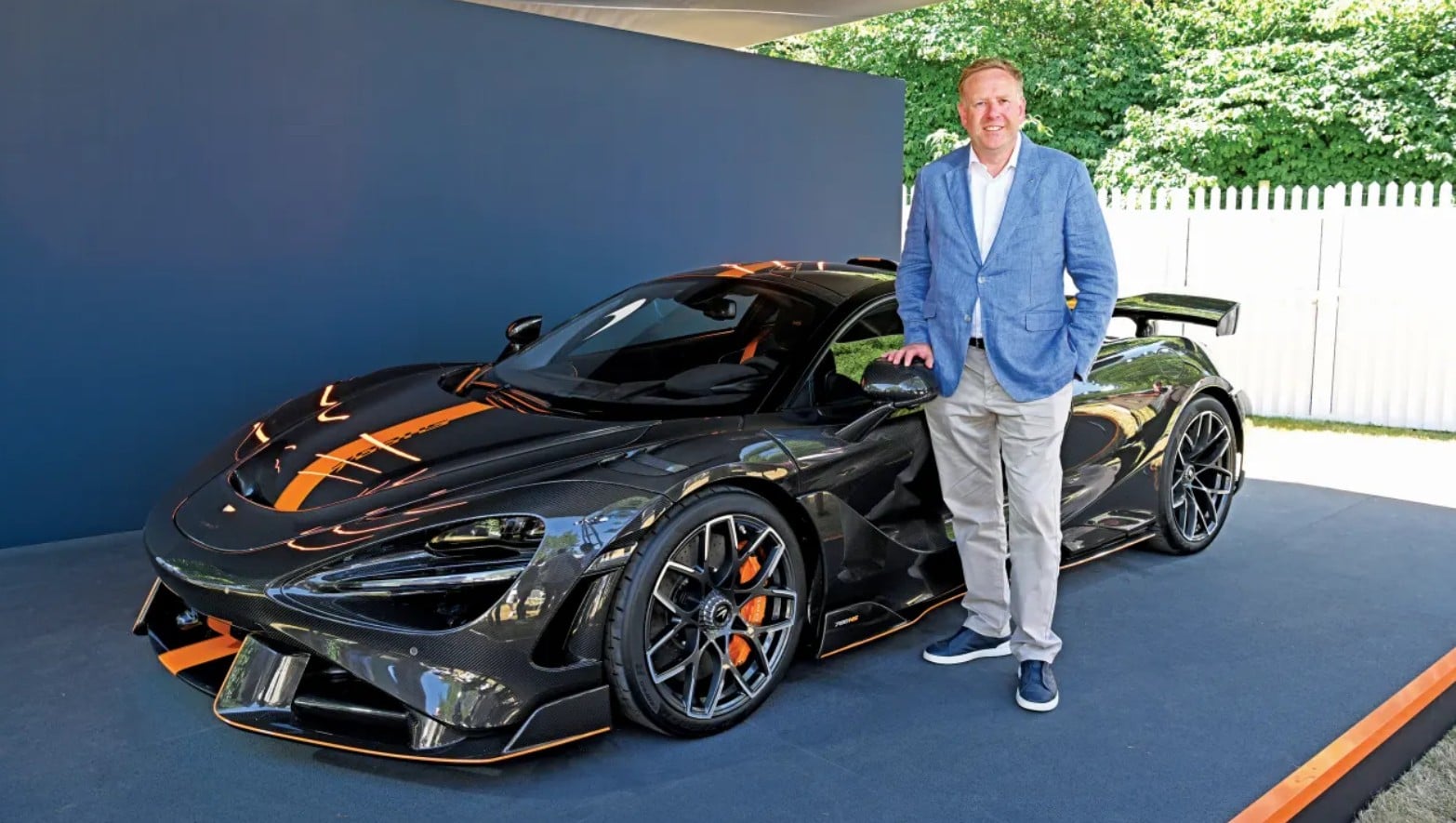 Man in a blue blazer stands beside a black carbon-fiber sports car with orange accents.