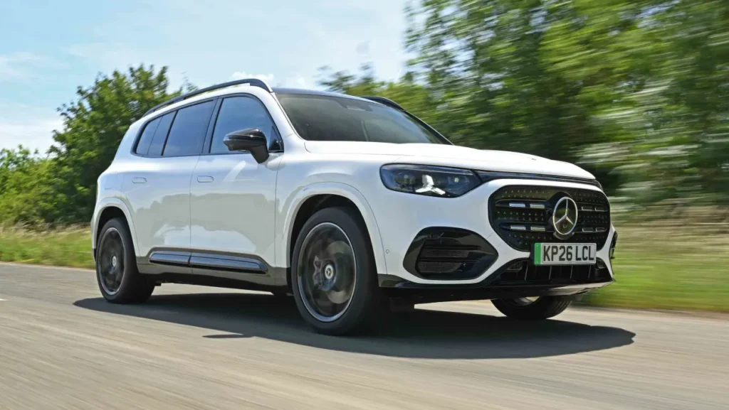 White Mercedes-Benz SUV driving on a tree-lined road, front grille visible and motion blur indicating speed.