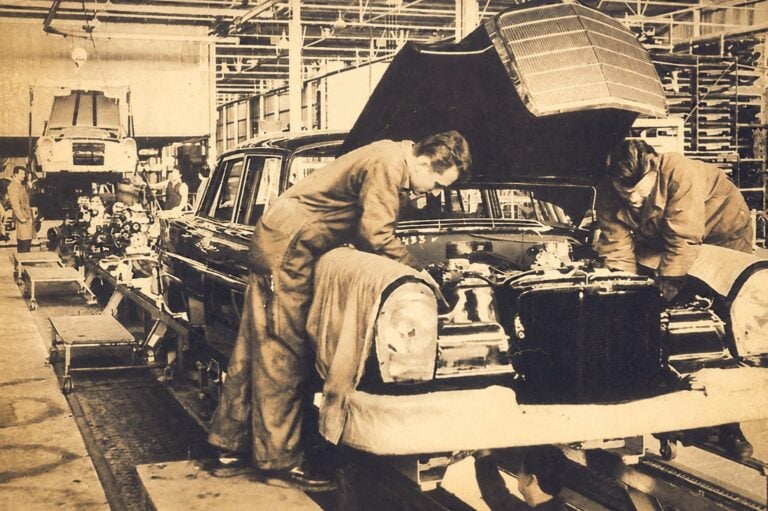 Workers assemble a vintage car on an early assembly line in a large factory, hood open and components exposed.