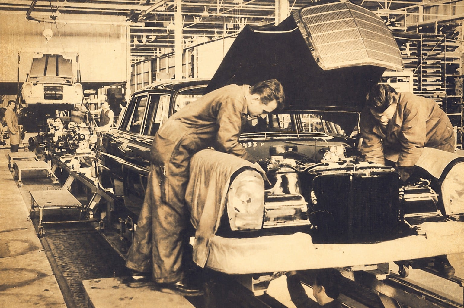 Workers assemble a vintage car on an early assembly line in a large factory, hood open and components exposed.