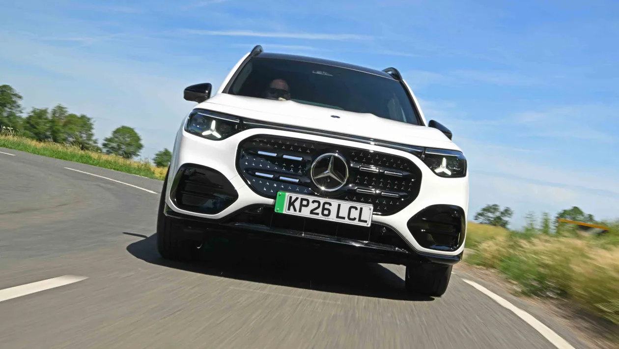 White Mercedes SUV driving on a curved road, angled toward the camera, with blue sky and green fields in the background.