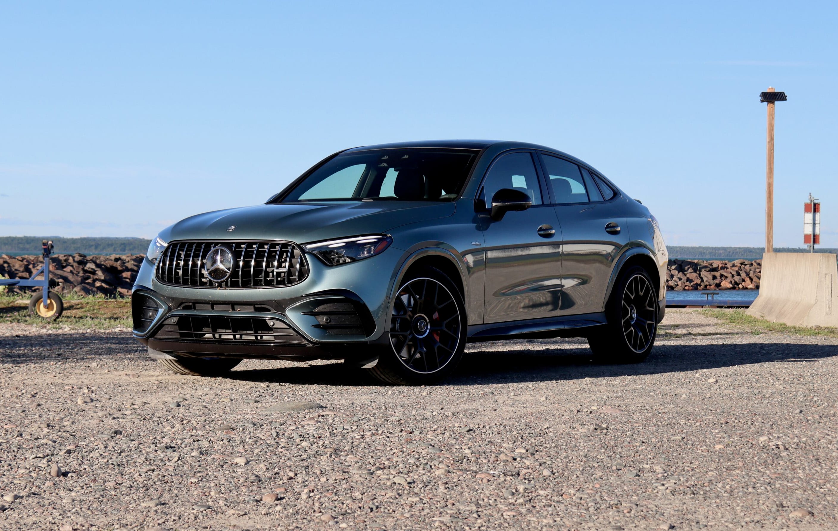 Blue Mercedes-Benz SUV parked on rocky ground by a harbor under a clear blue sky. (Front three-quarter view)