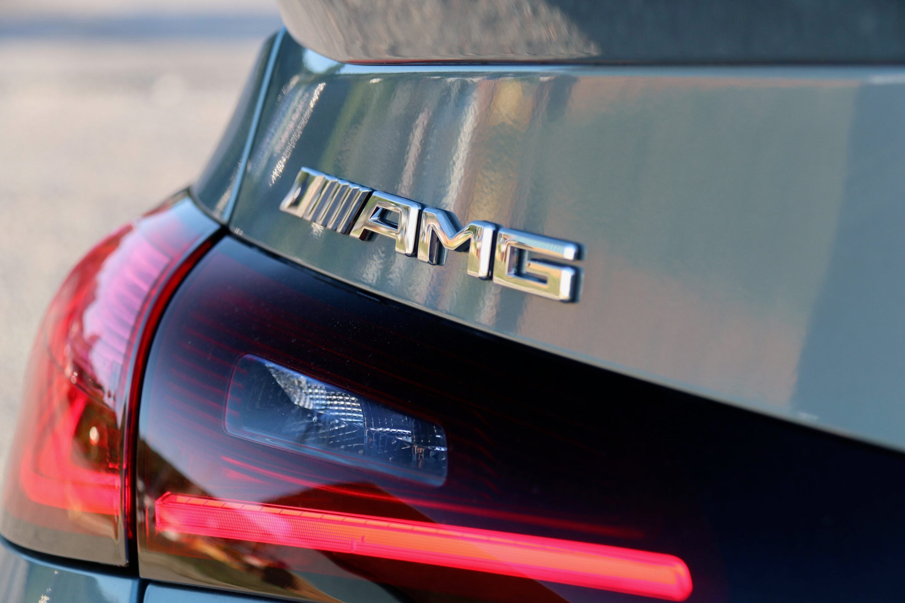 Close-up of a chrome badge on the rear of a dark car, with a red taillight visible below.