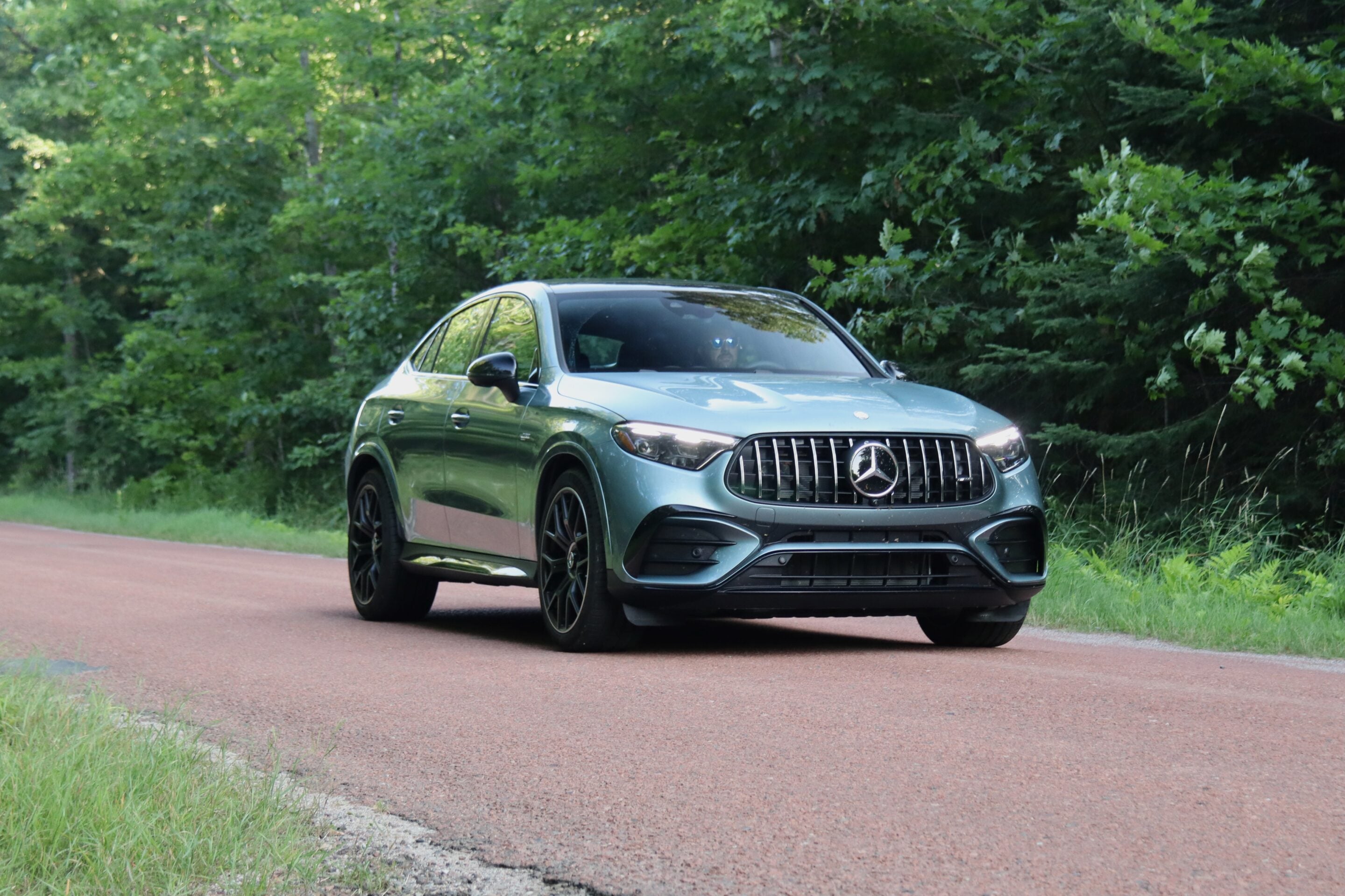 Silver-green Mercedes SUV driving on a red paved road with lush green trees and grasses on both sides, front grille visible.