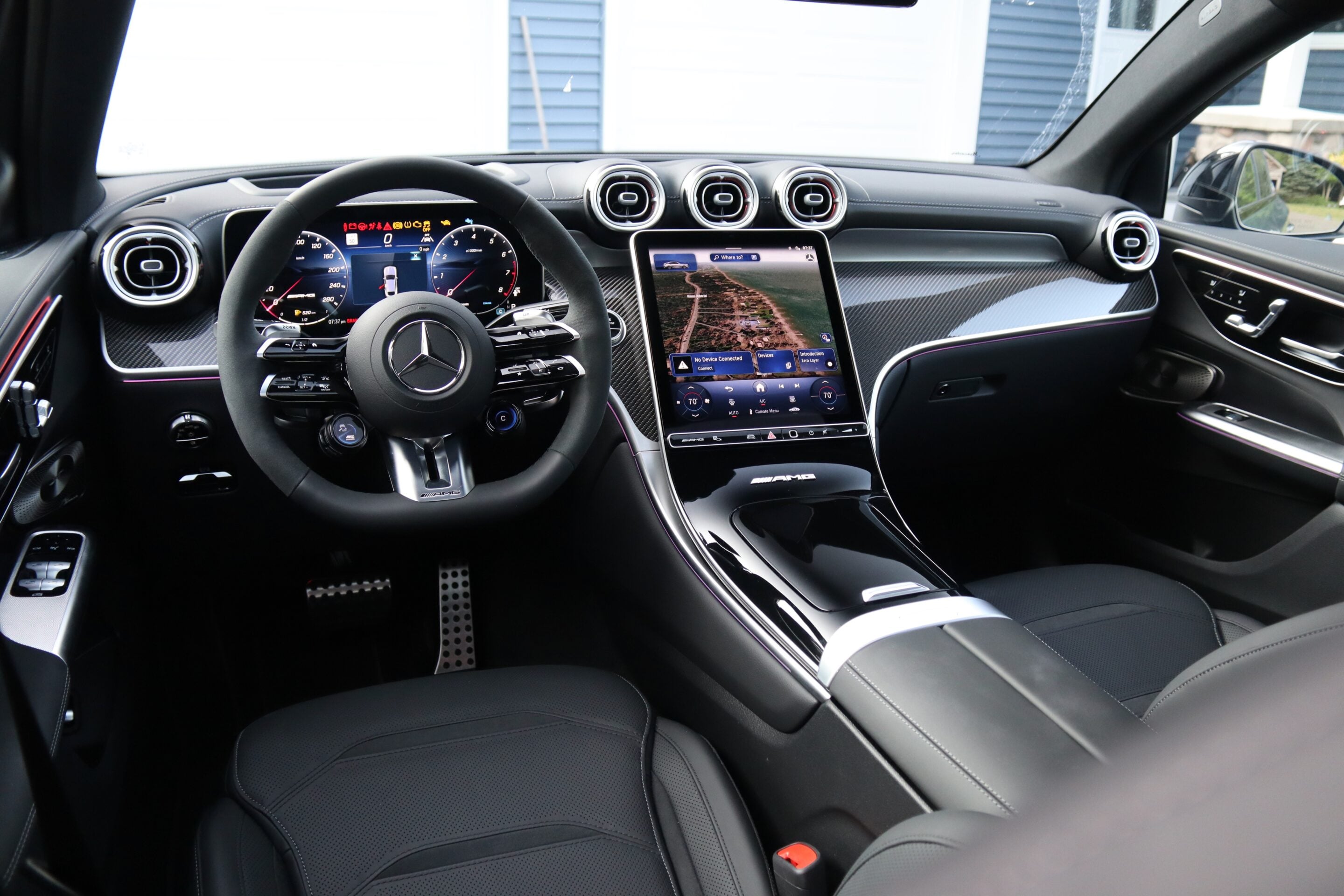Luxury car interior: steering wheel with Mercedes emblem, digital instrument cluster, and a large vertical touchscreen in the center surrounded by carbon-fiber trim and ambient lighting.