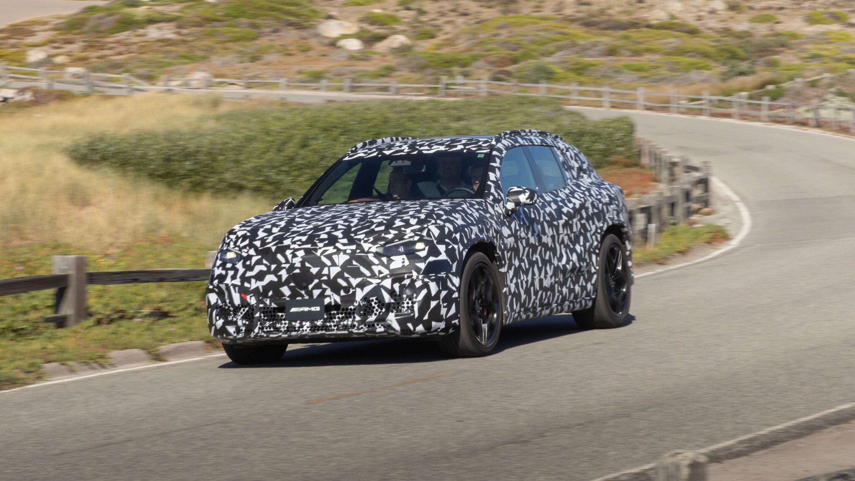 Camouflaged prototype SUV with geometric black-and-white wrap driving on a winding rural road, two occupants visible inside.