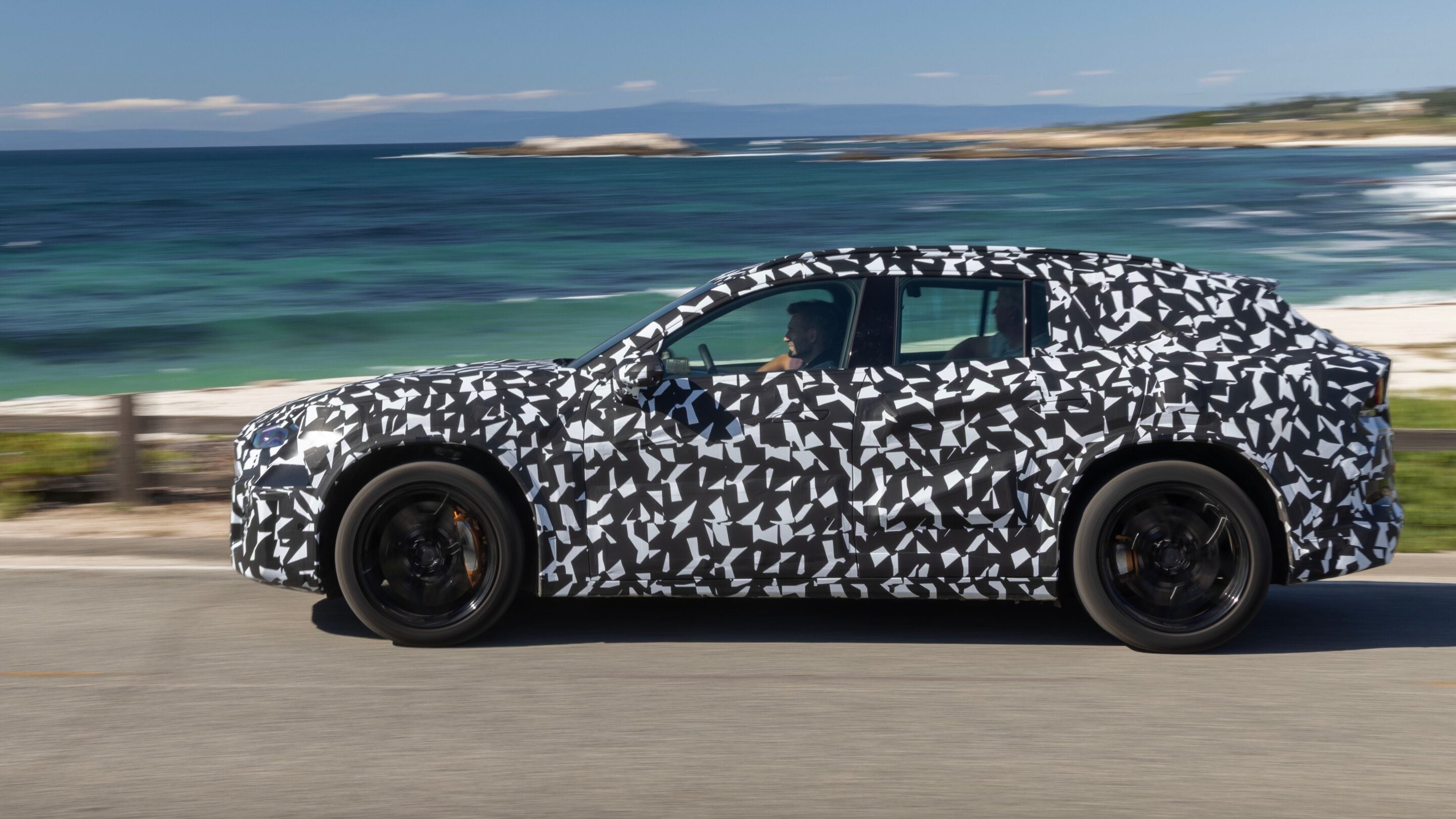 Side view of a camouflaged test car speeding along a coastal highway with the sea in the background.