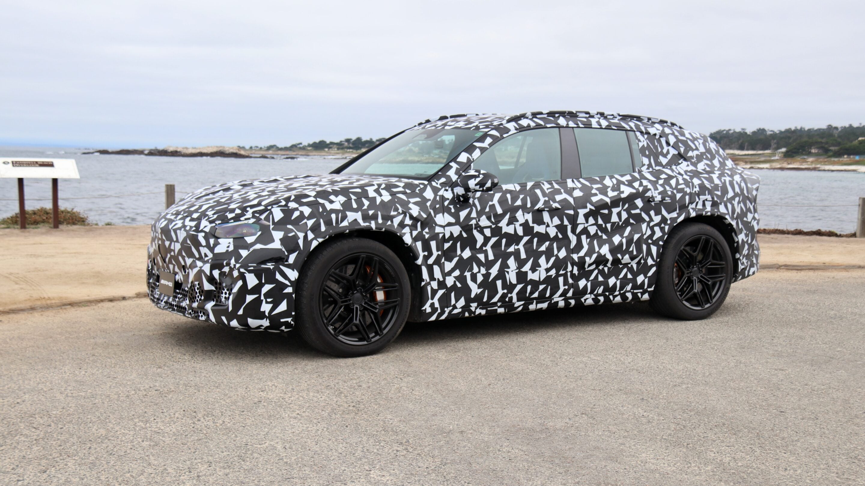 Camouflaged prototype sports car with black-and-white geometric wrap parked along a coastal road by the sea.