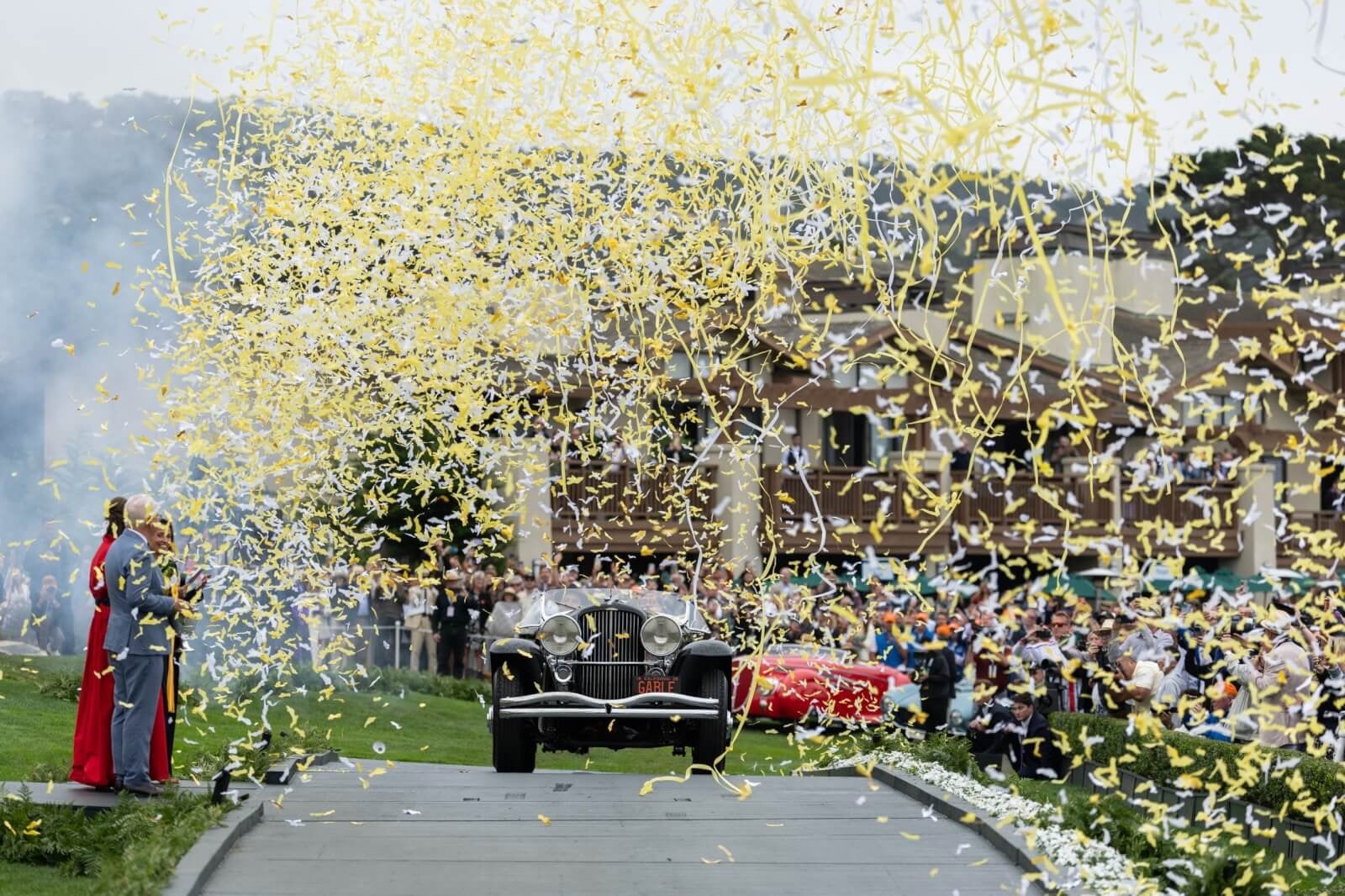 Vintage car driving through a shower of yellow and white confetti as spectators cheer along a parade route.