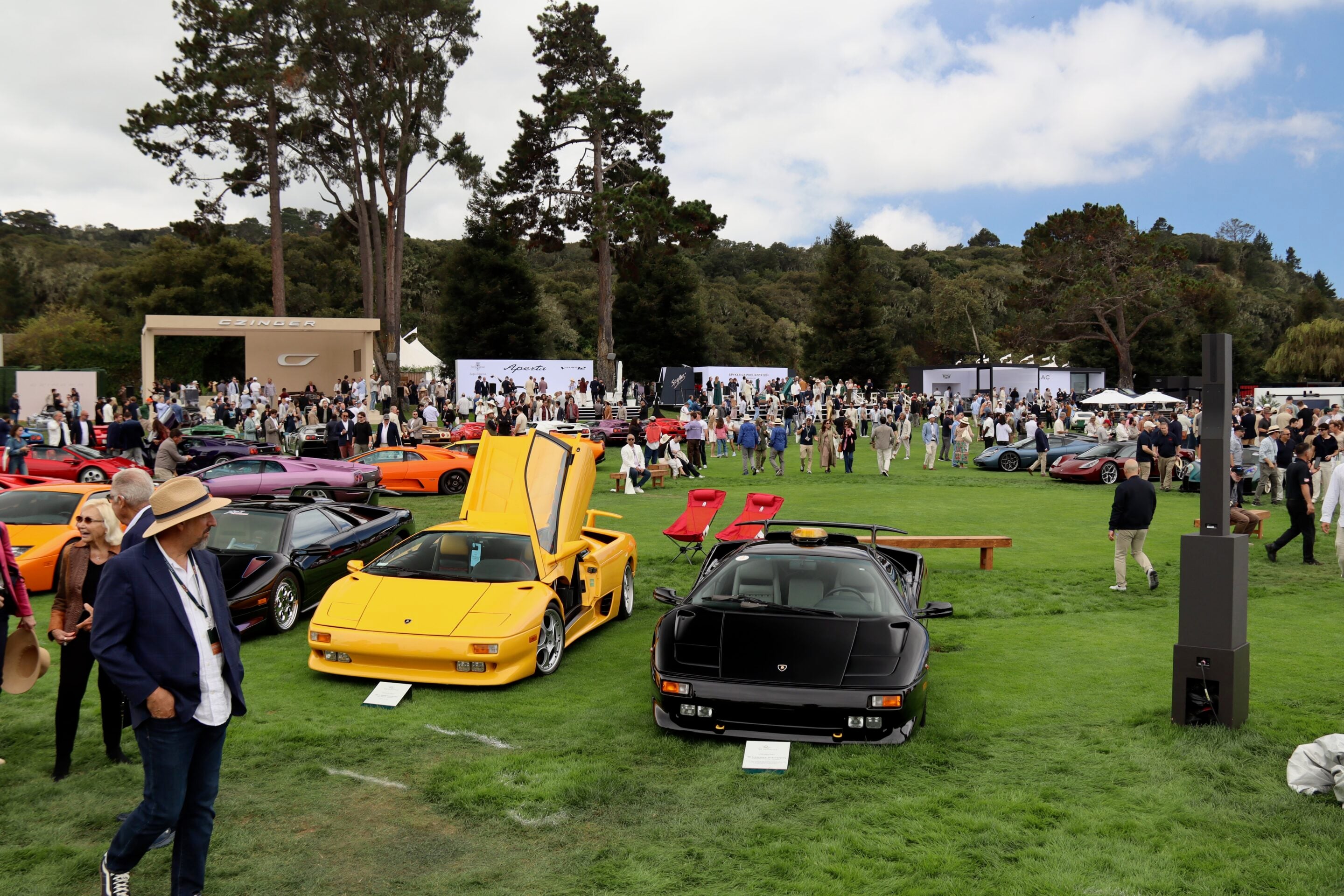 Outdoor car show on a grassy field with colorful sports cars and a crowd under a blue sky with clouds.