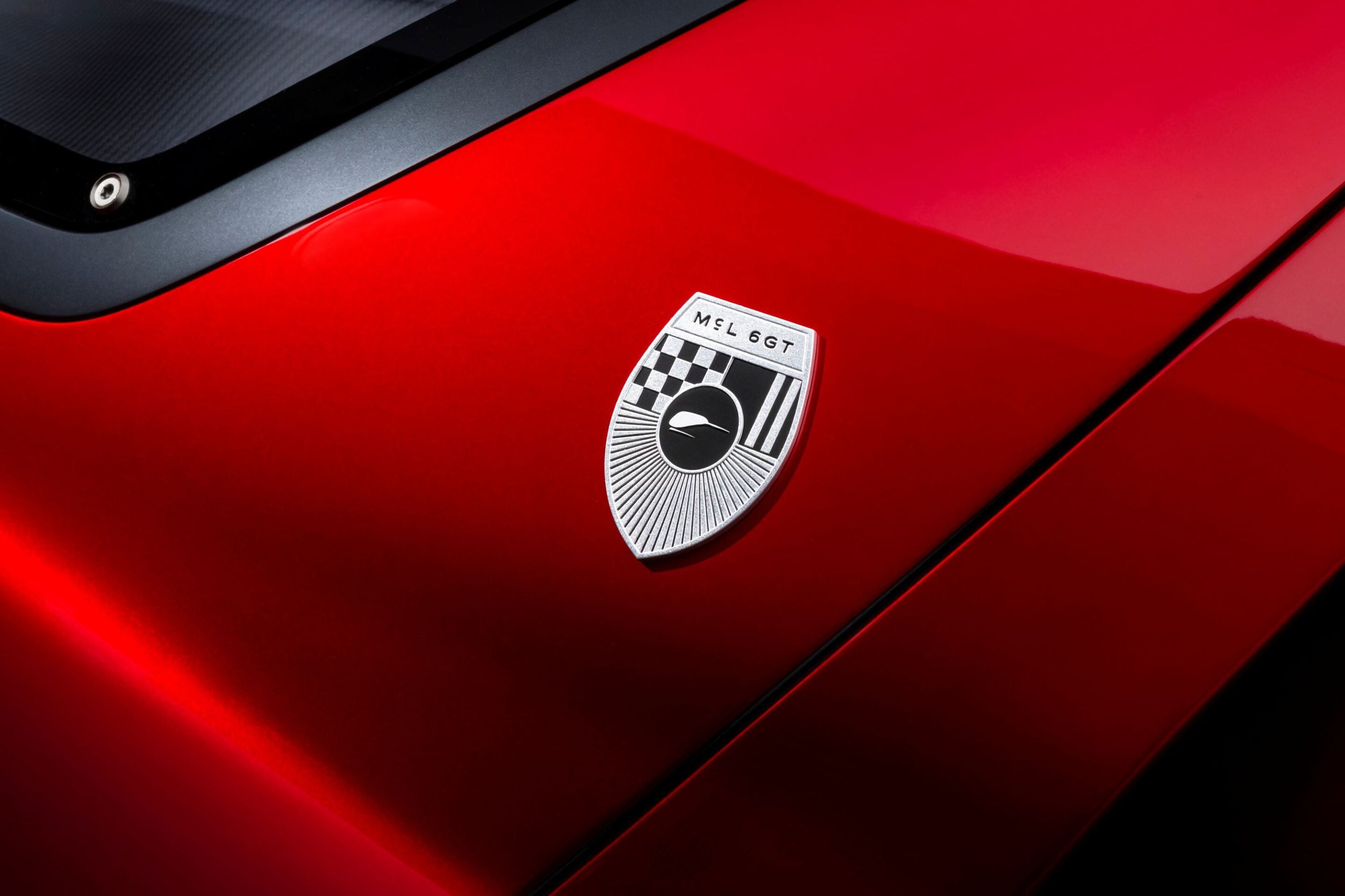 Close-up of a red car hood featuring a chrome badge with a checkered emblem and the text 'MEL 6GT' near the center-right edge.