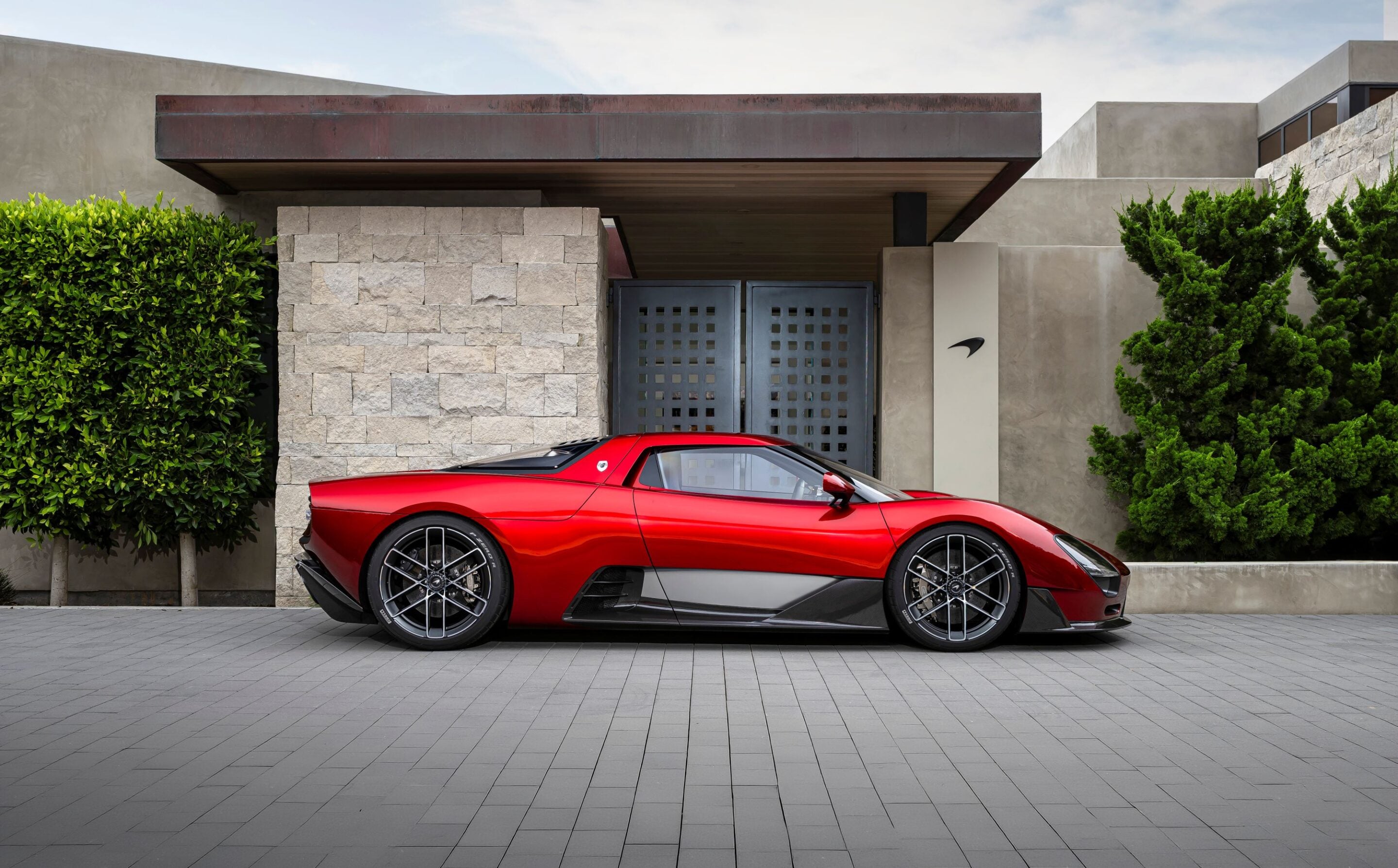 Red sports car parked in front of a modern gray stone-and-concrete house with green hedges on both sides.