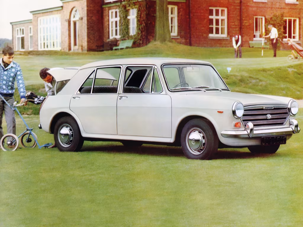 Vintage white four-door sedan parked on a well-manicured golf course with its trunk open and people nearby, brick clubhouse in the background.