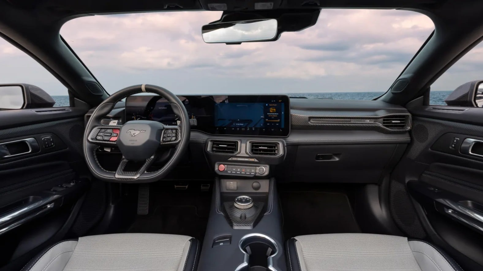 Modern car interior with a large center touchscreen, steering wheel, and carbon-fiber trim, ocean visible through the windshield at the horizon amidst a cloudy sky.