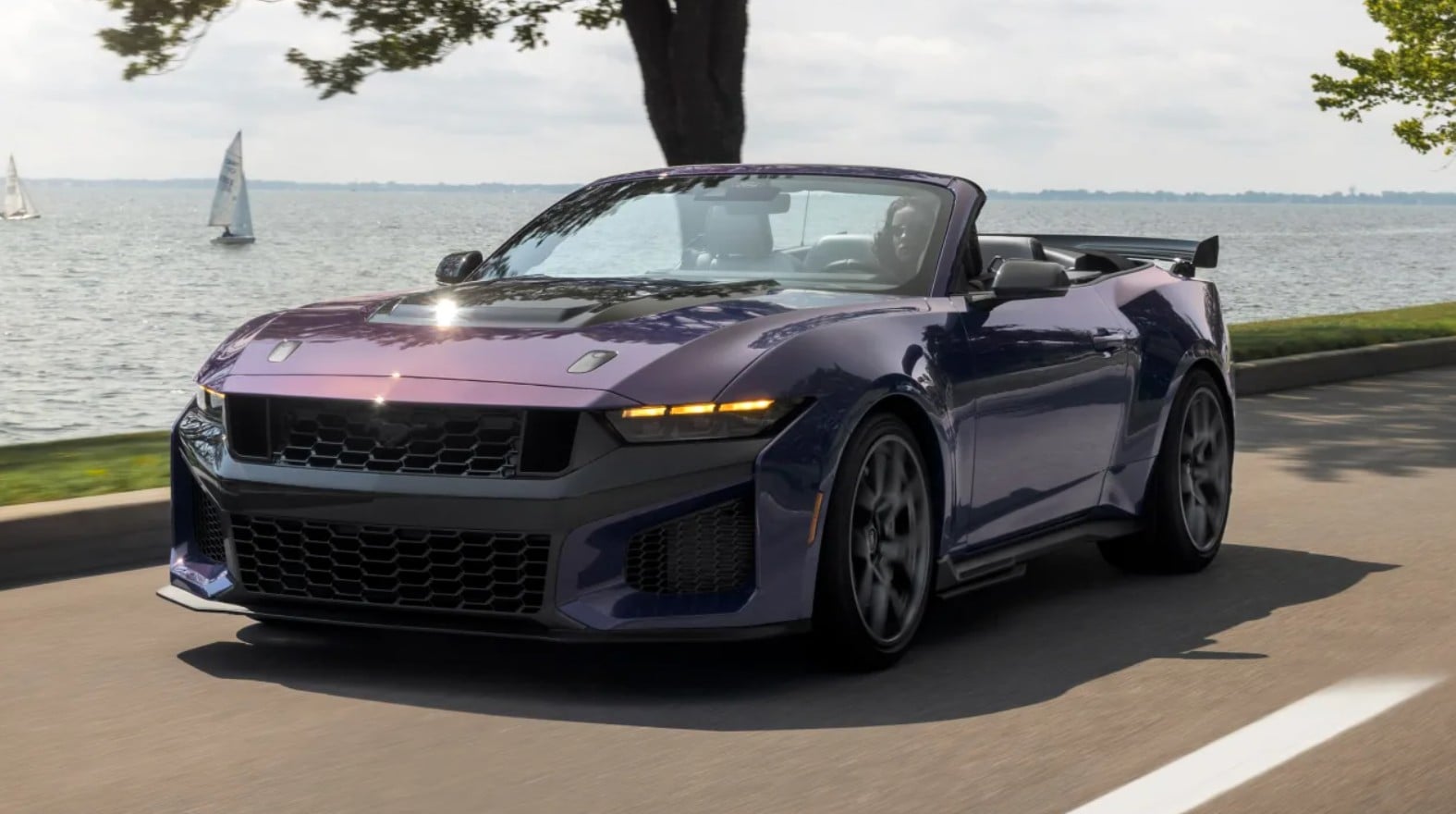 Purple convertible sports car driving along a lakeside road, top down, with water and sailboats in the background.