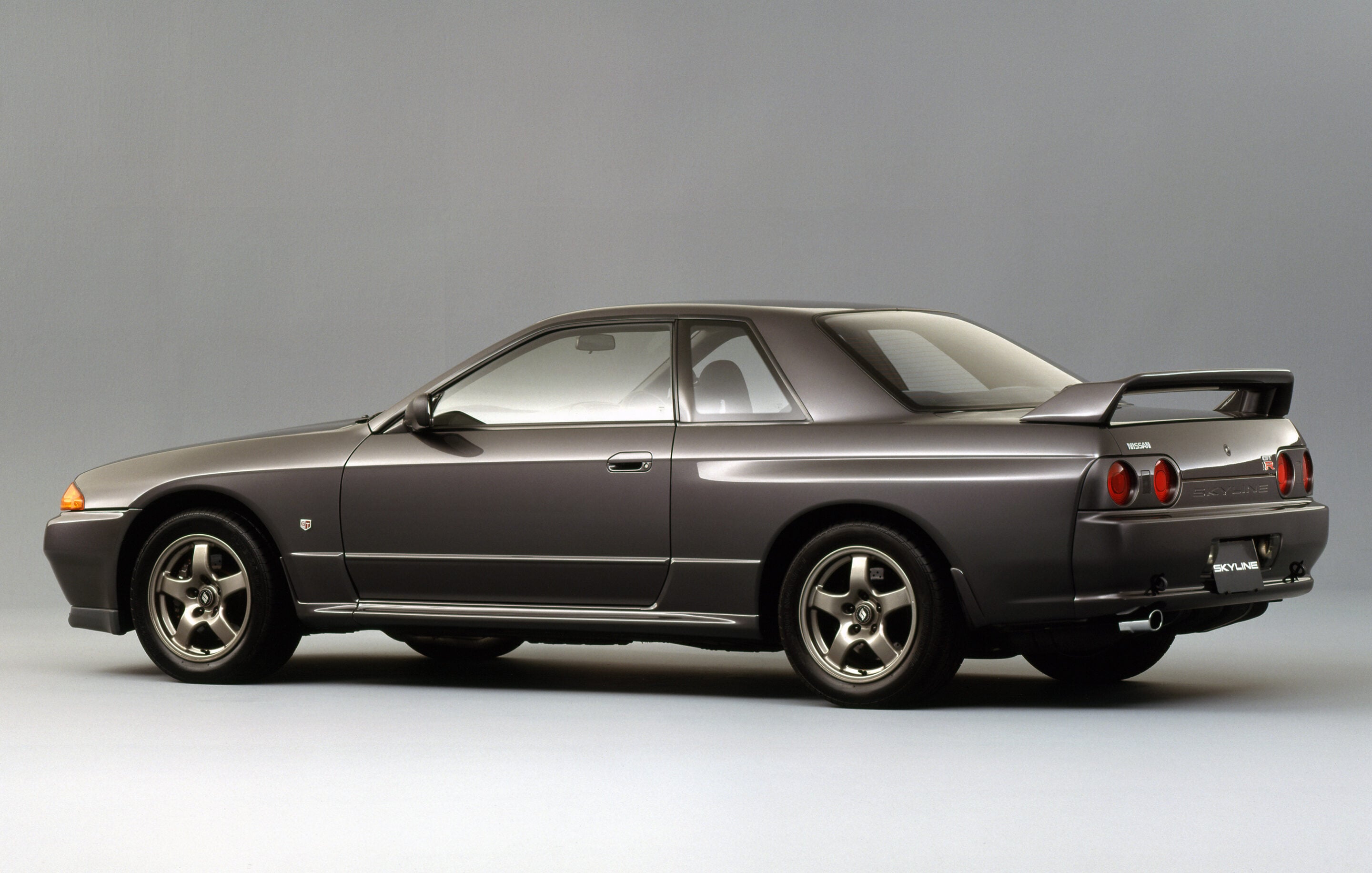 Rear three-quarter view of a metallic gray Nissan Skyline GT-R (R34) in a studio setting with a prominent rear wing.