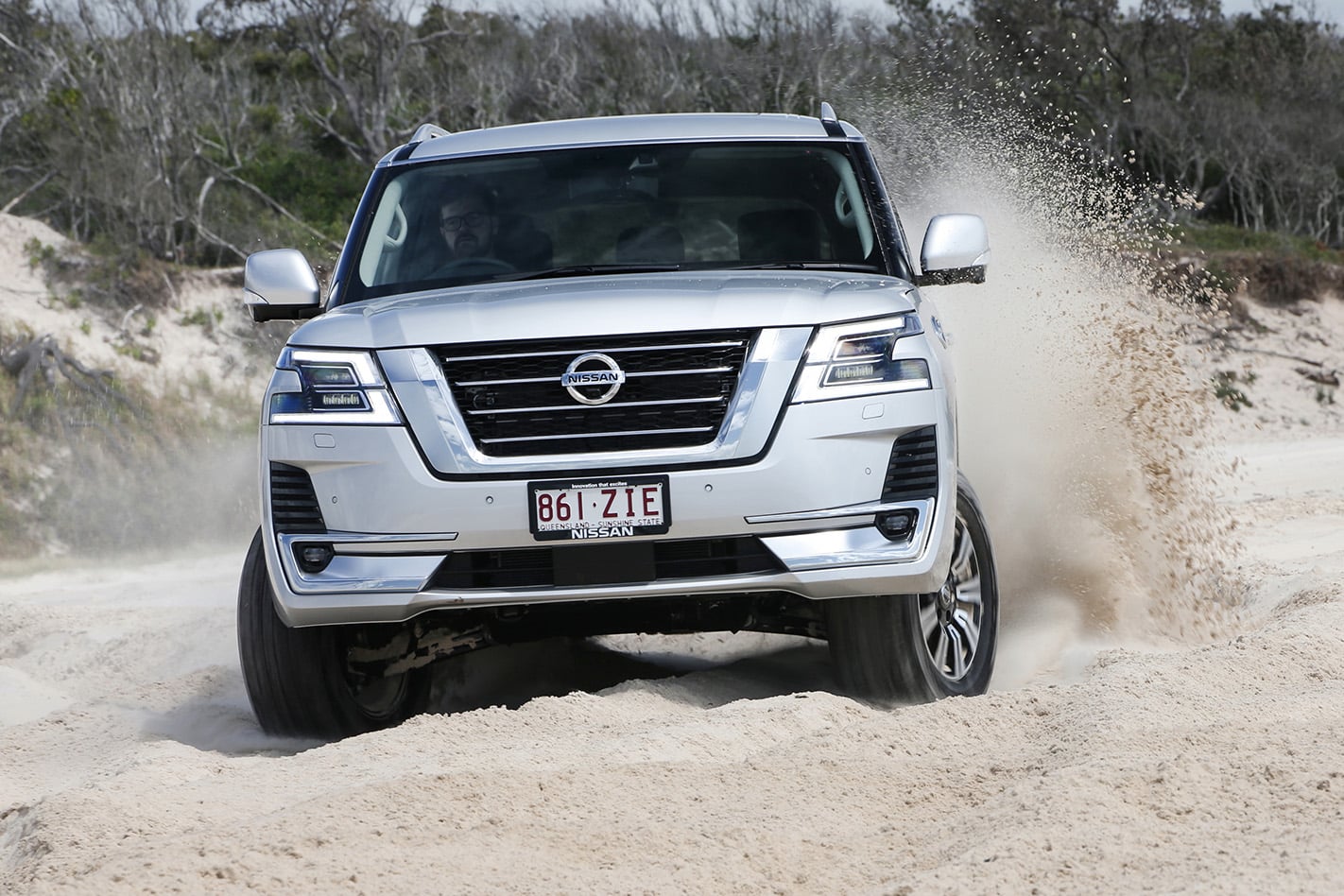 Silver Nissan SUV kicking up dirt while turning on a sandy off-road trail, headlights on, driver visible through windshield