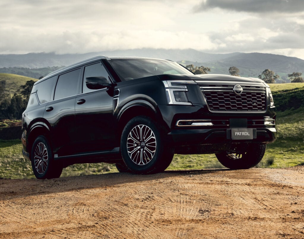2026 Nissan Y63 Patrol finished in black in a static front three-quarter exterior view parked on a dirt hill with rolling green hills, distant mountains and a cloudy sky in the background.