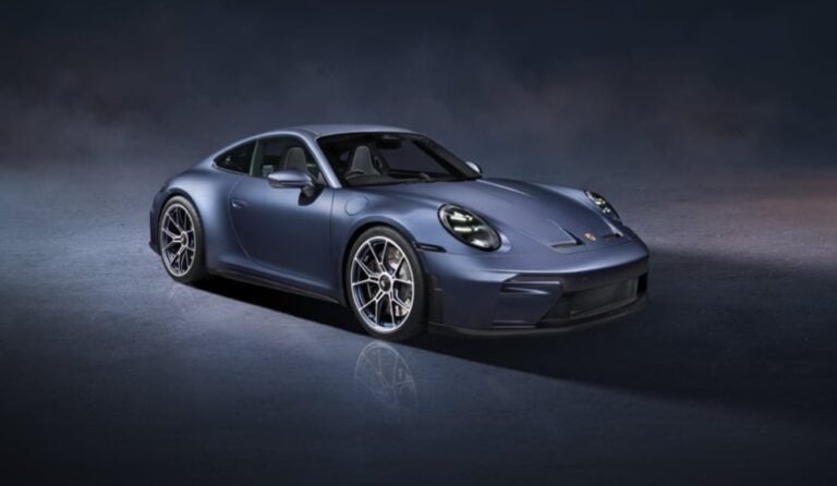 Three-quarter front view of a blue Porsche sports car parked on a dark, misty backdrop, with reflections on the ground