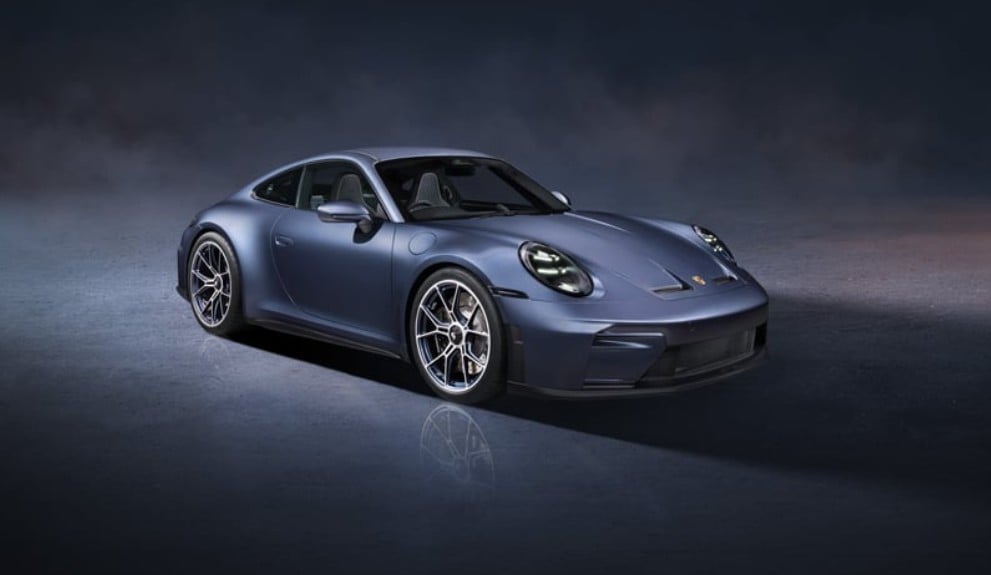 Three-quarter front view of a blue Porsche sports car parked on a dark, misty backdrop, with reflections on the ground
