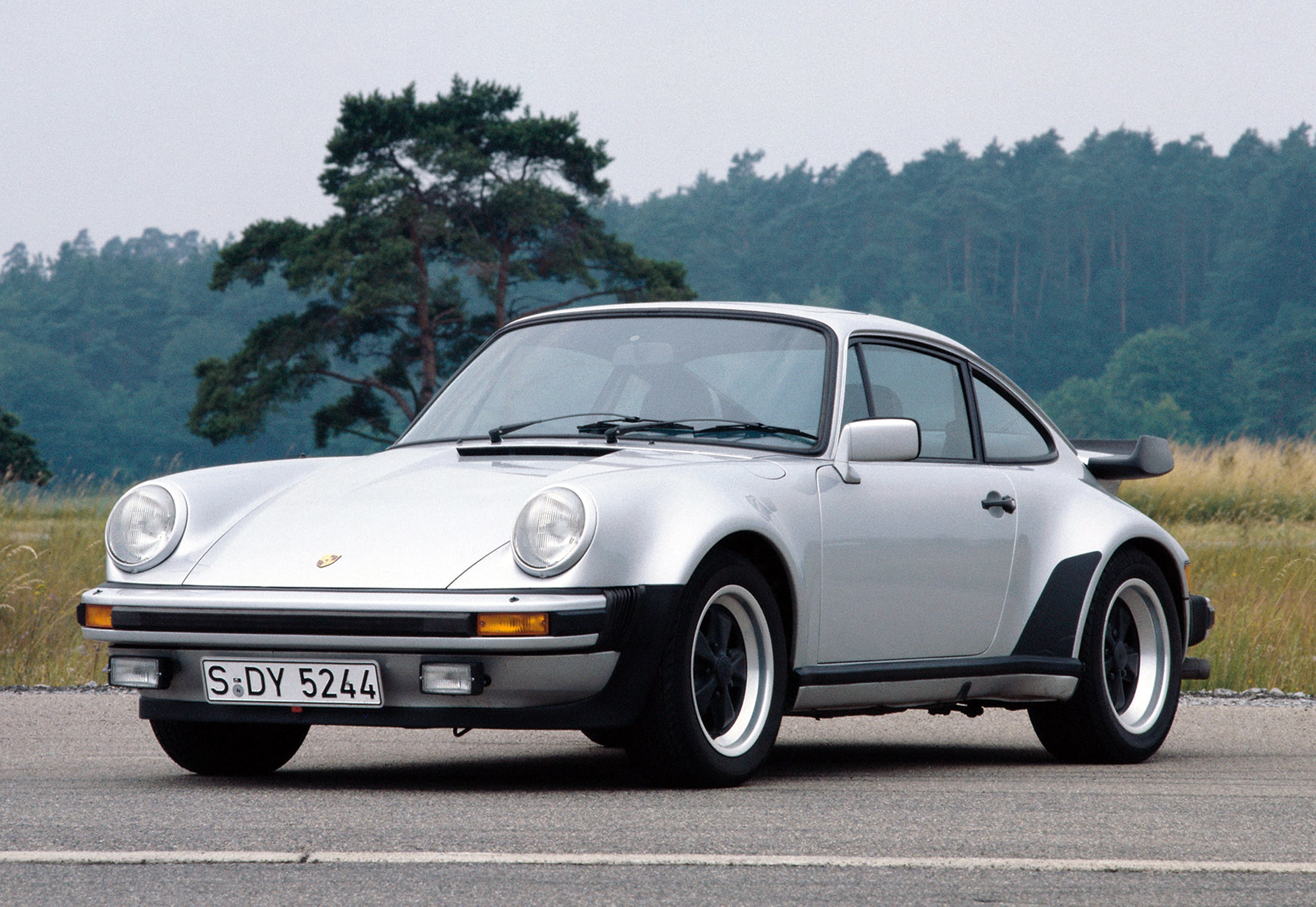 Front view of a white Porsche sports car parked on a rural road with a forest in the background, showcasing its rounded headlights and bumper.