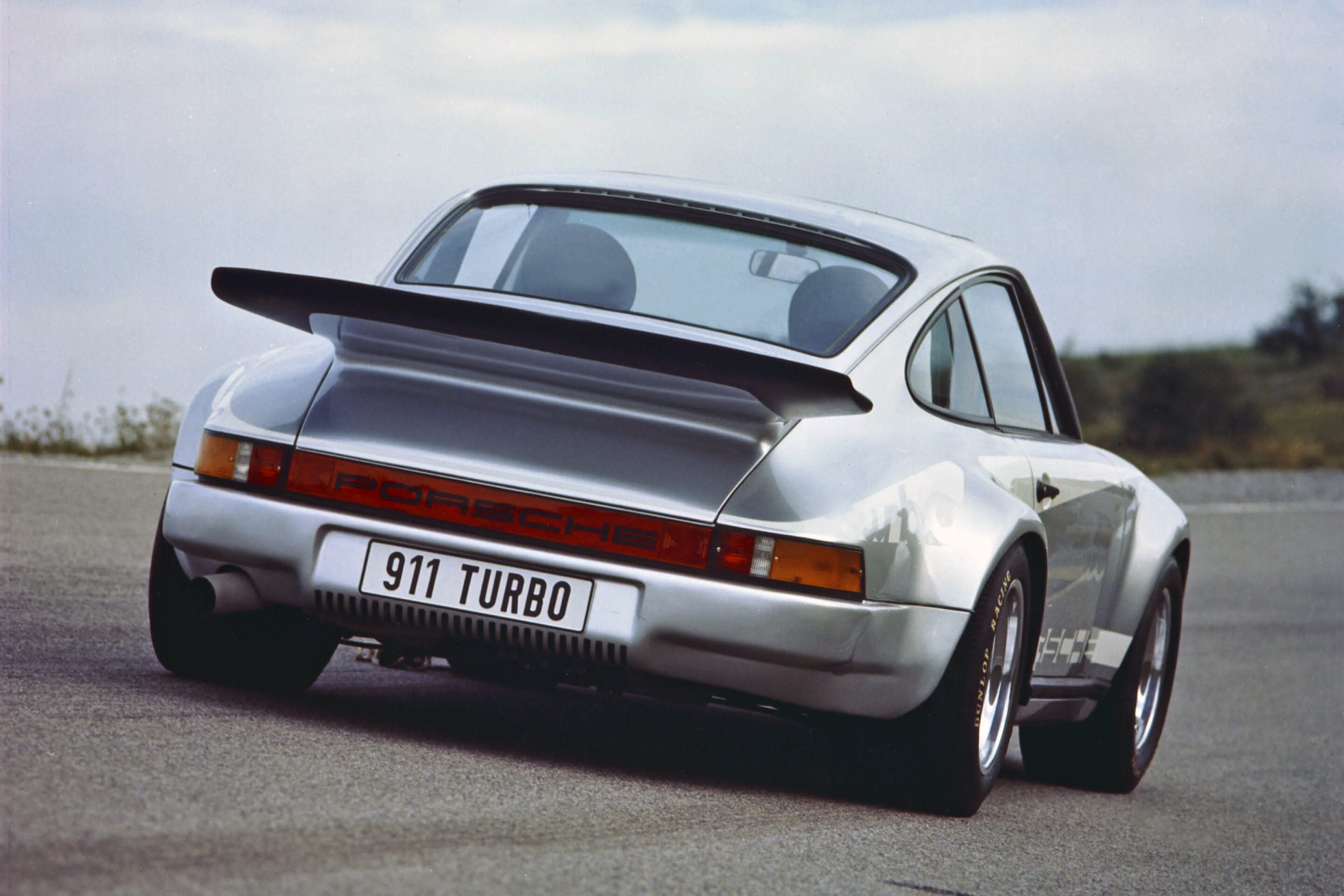 Rear three-quarter view of a silver Porsche 911 Turbo on a road, featuring a large black rear wing and red taillights across the back.