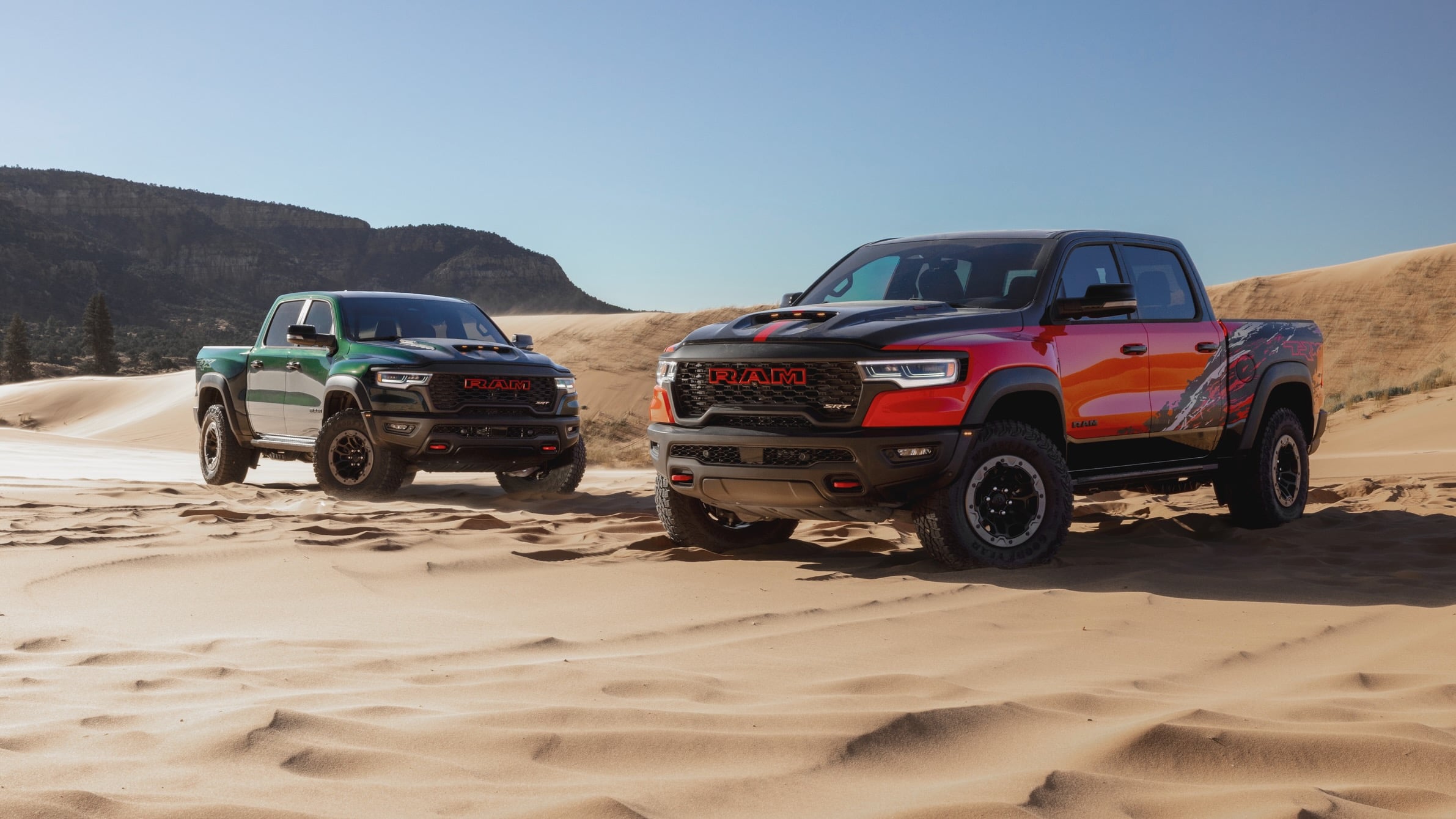 Two RAM pickup trucks parked on sandy desert dunes with a rocky hill in the background, under a clear blue sky.