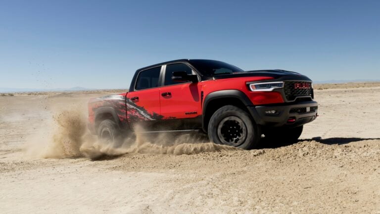 Red RAM pickup kicking up dust in a sandy desert with clear blue sky, showing off-road tires and graphics on the side panel.