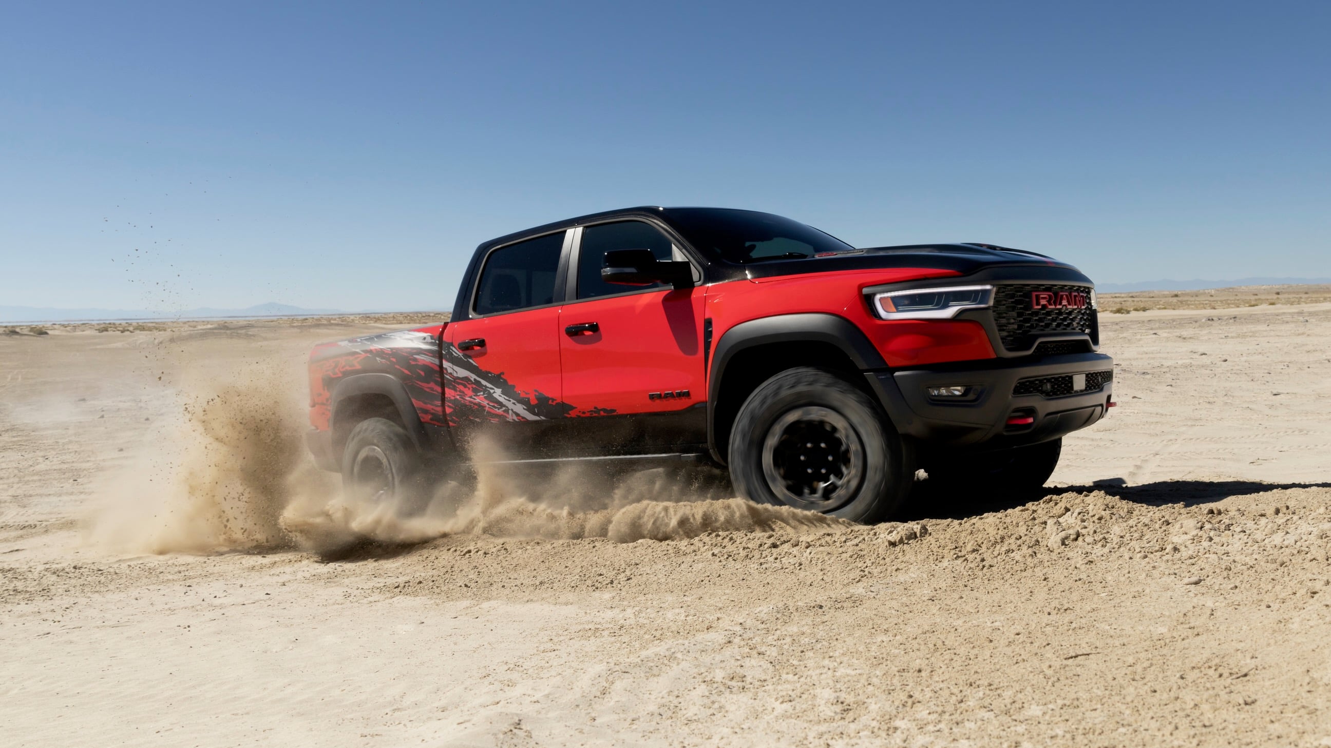 Red RAM pickup kicking up dust in a sandy desert with clear blue sky, showing off-road tires and graphics on the side panel.