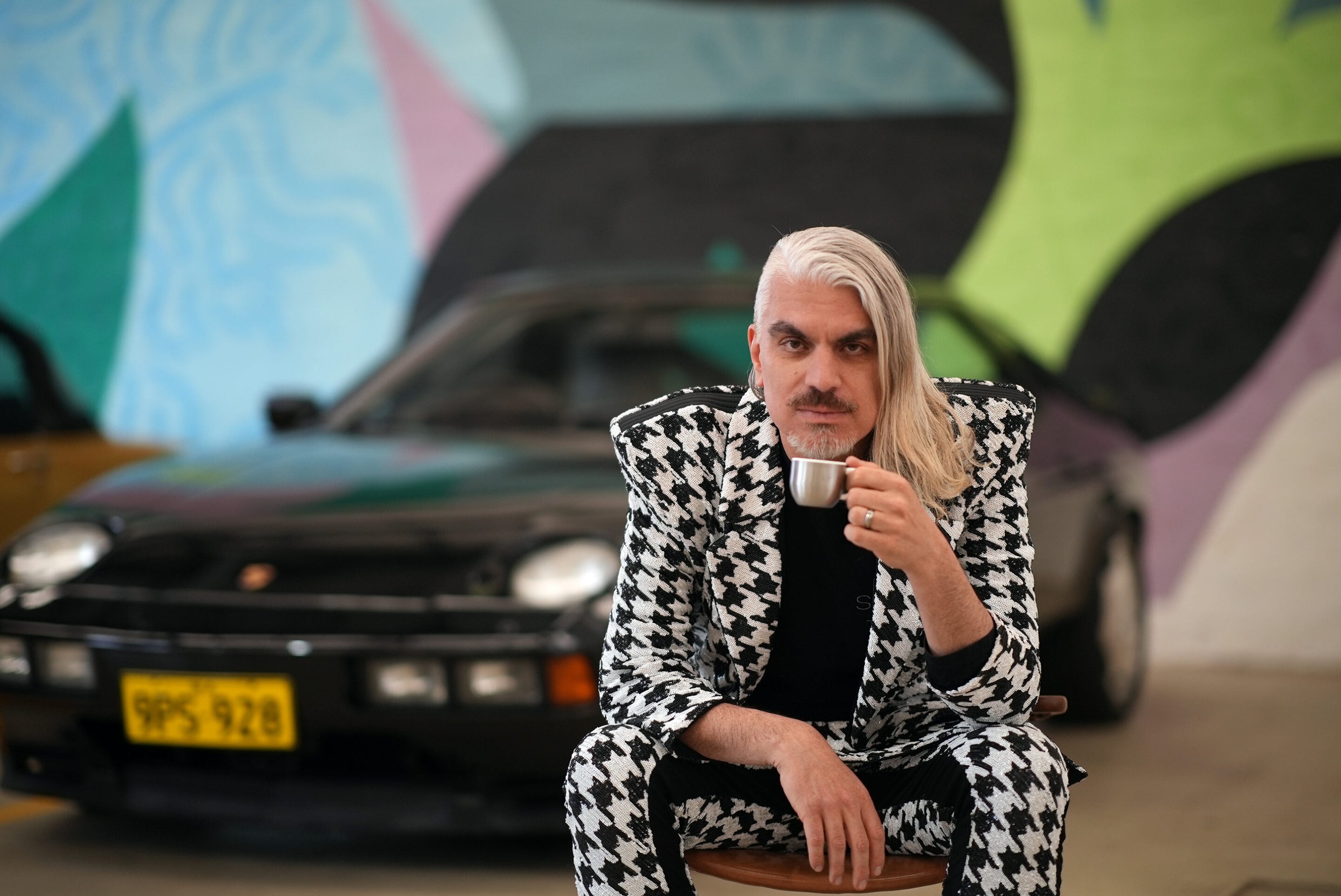 Person with silver-blonde hair and a light beard sits on a stool, wearing a black-and-white houndstooth suit and holding a metal cup, with a graffiti backdrop and a car behind.