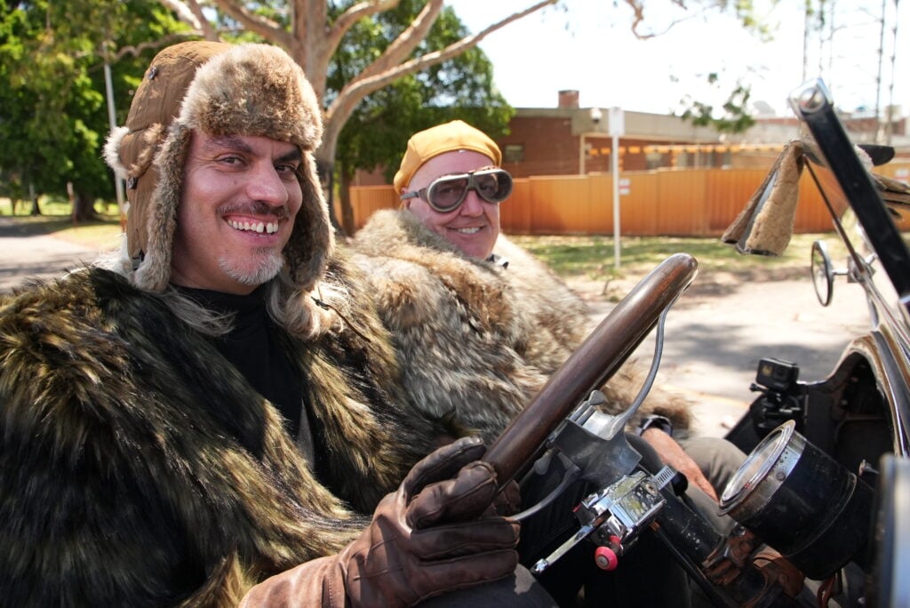 Two men in fur coats and aviator hats sit in a vintage convertible, smiling at the camera.