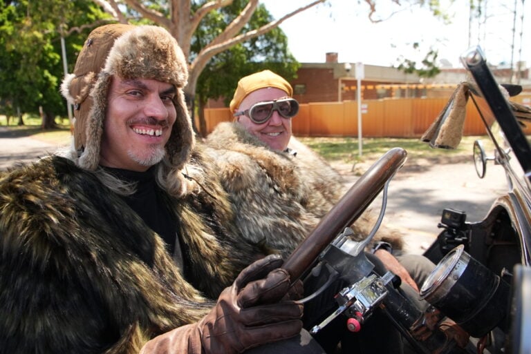 Two men in fur coats and aviator hats sit in a vintage convertible, smiling at the camera.