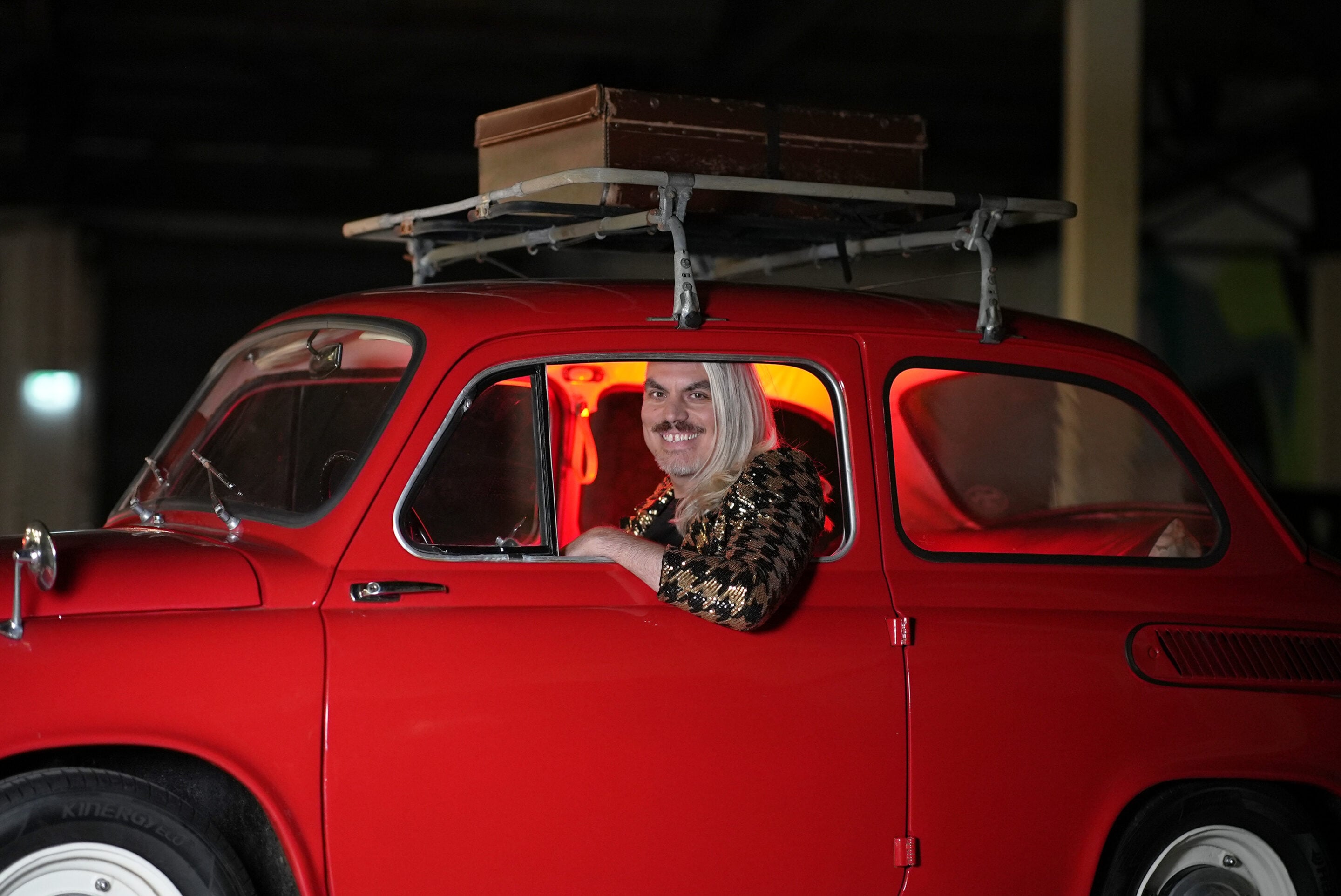 Man with a long blonde wig and a sequined outfit sits in the driver's seat of a red vintage car at night, with a wooden box on a roof rack above him.