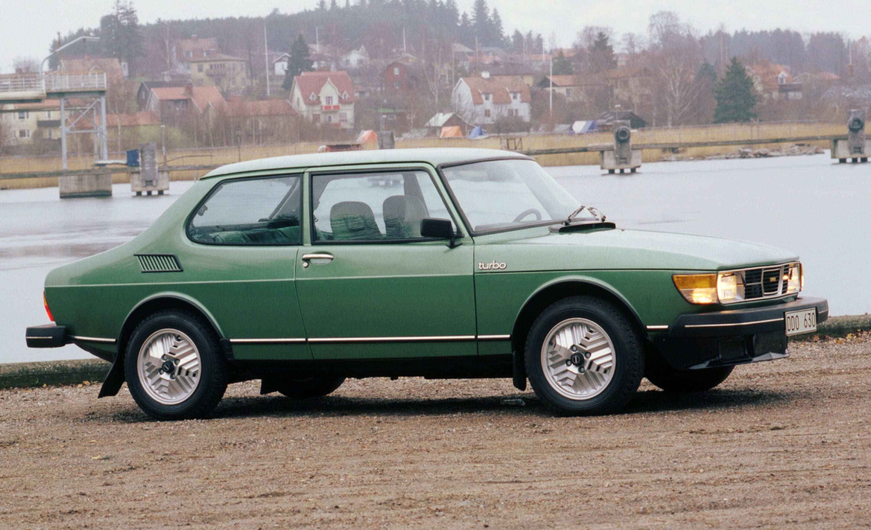 Green vintage sedan parked by a calm waterfront with a small town and hills in the background, showing a classic two-door car profile.