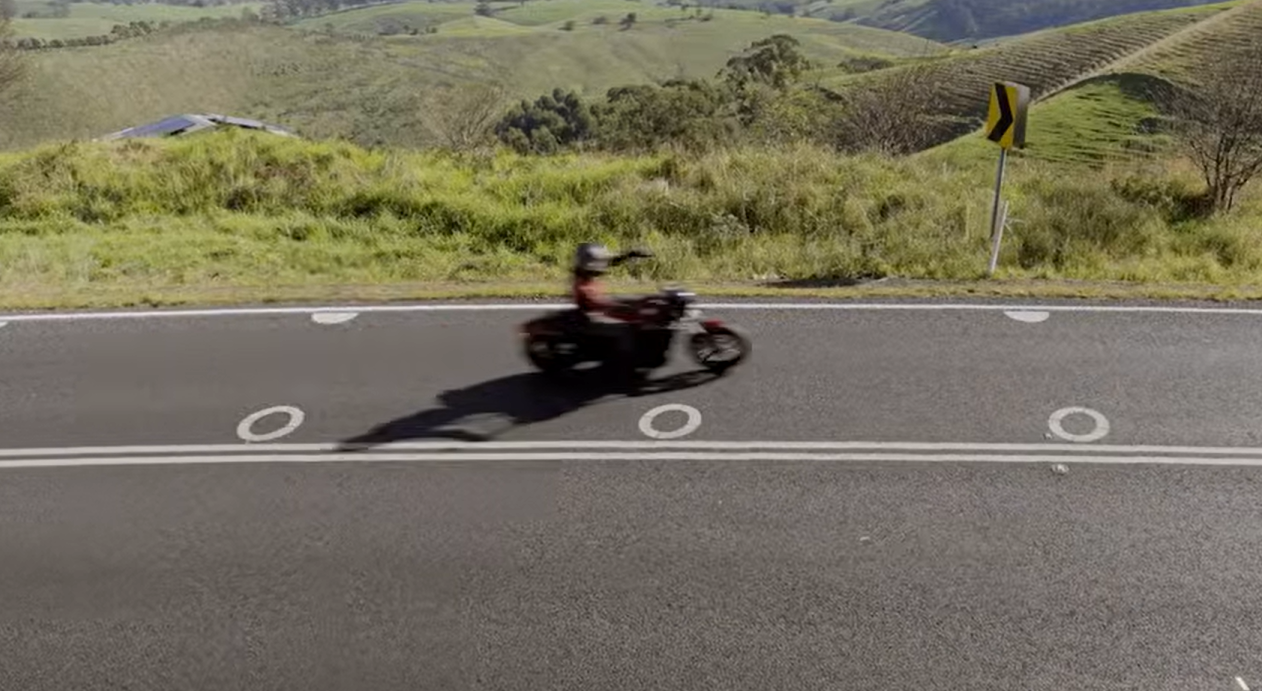 Motorcyclist riding a motorcycle on a two-lane rural road with a curve ahead sign and green hills in the background.