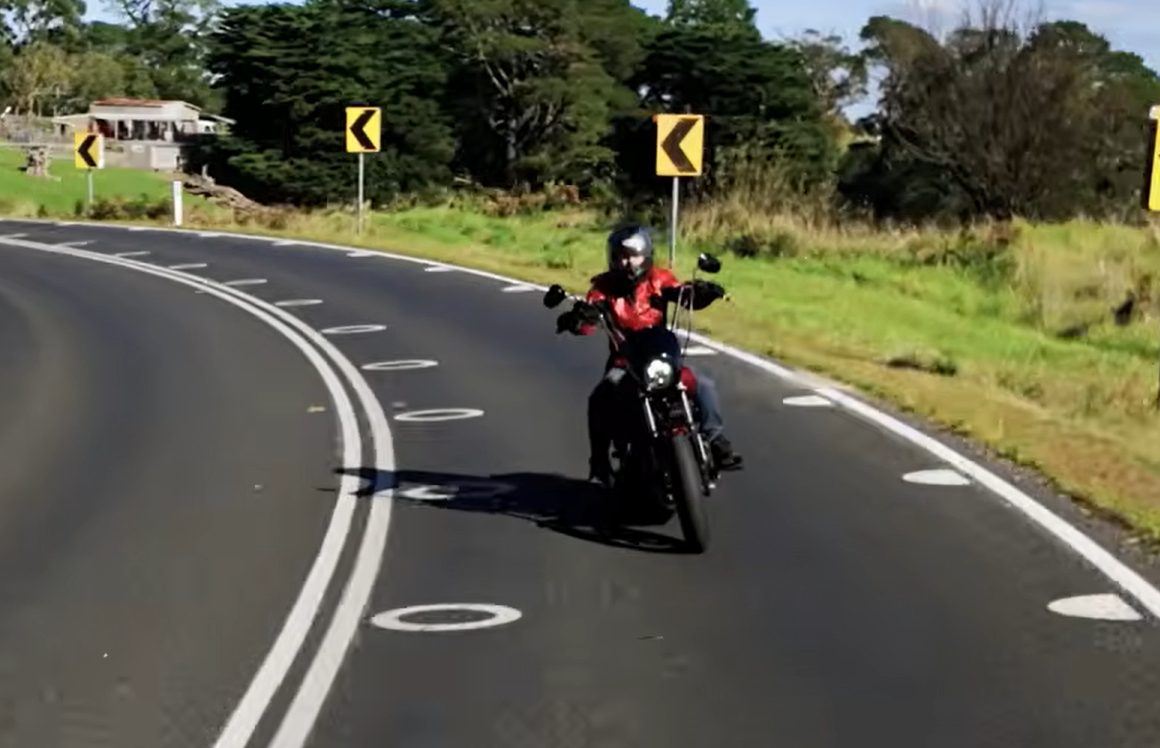Motorcyclist wearing a red jacket rides a black motorcycle around a left-hand curve on a rural road with chevron warning signs ahead.